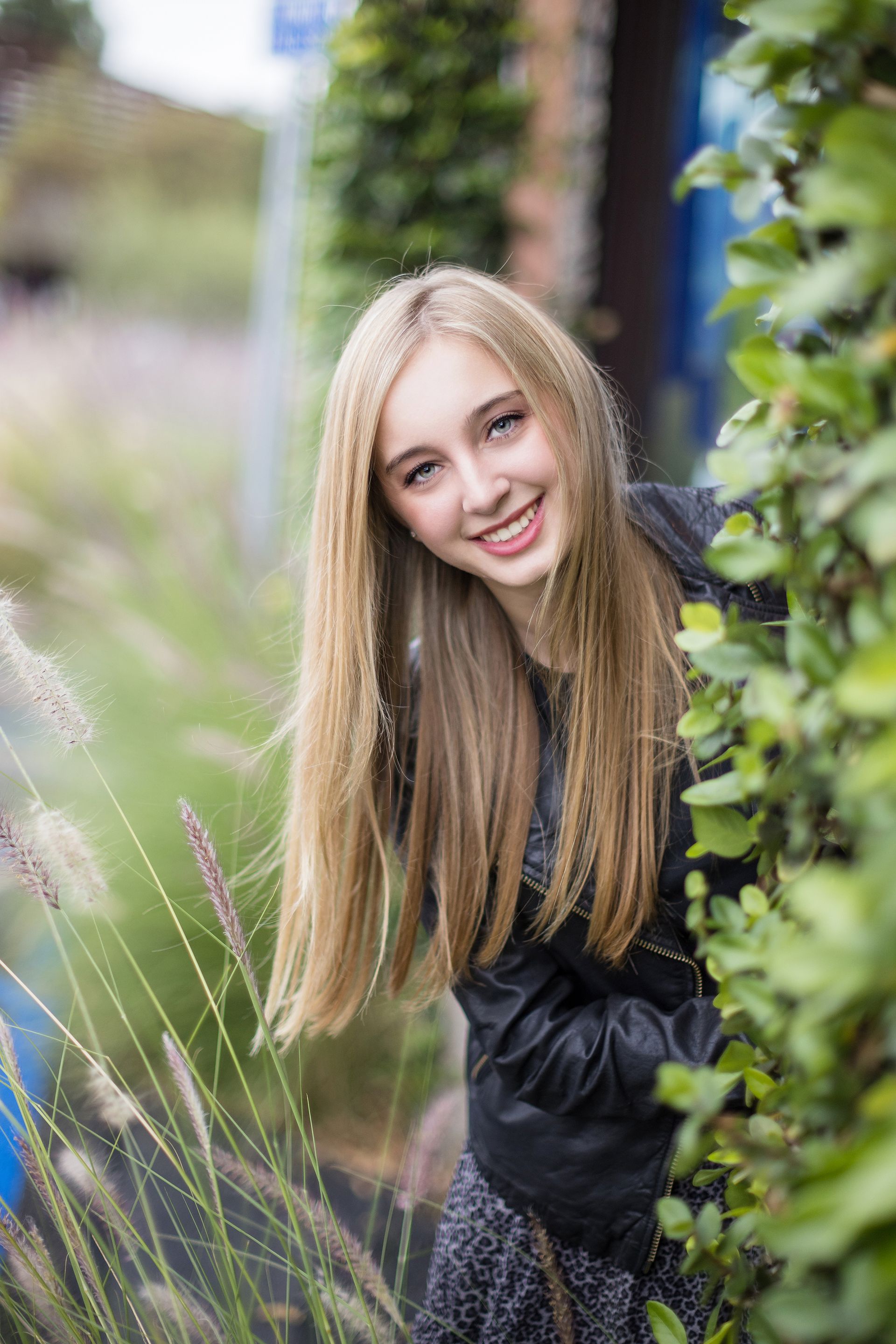 A blonde high school senior girl smiles brightly while leaning through greenery