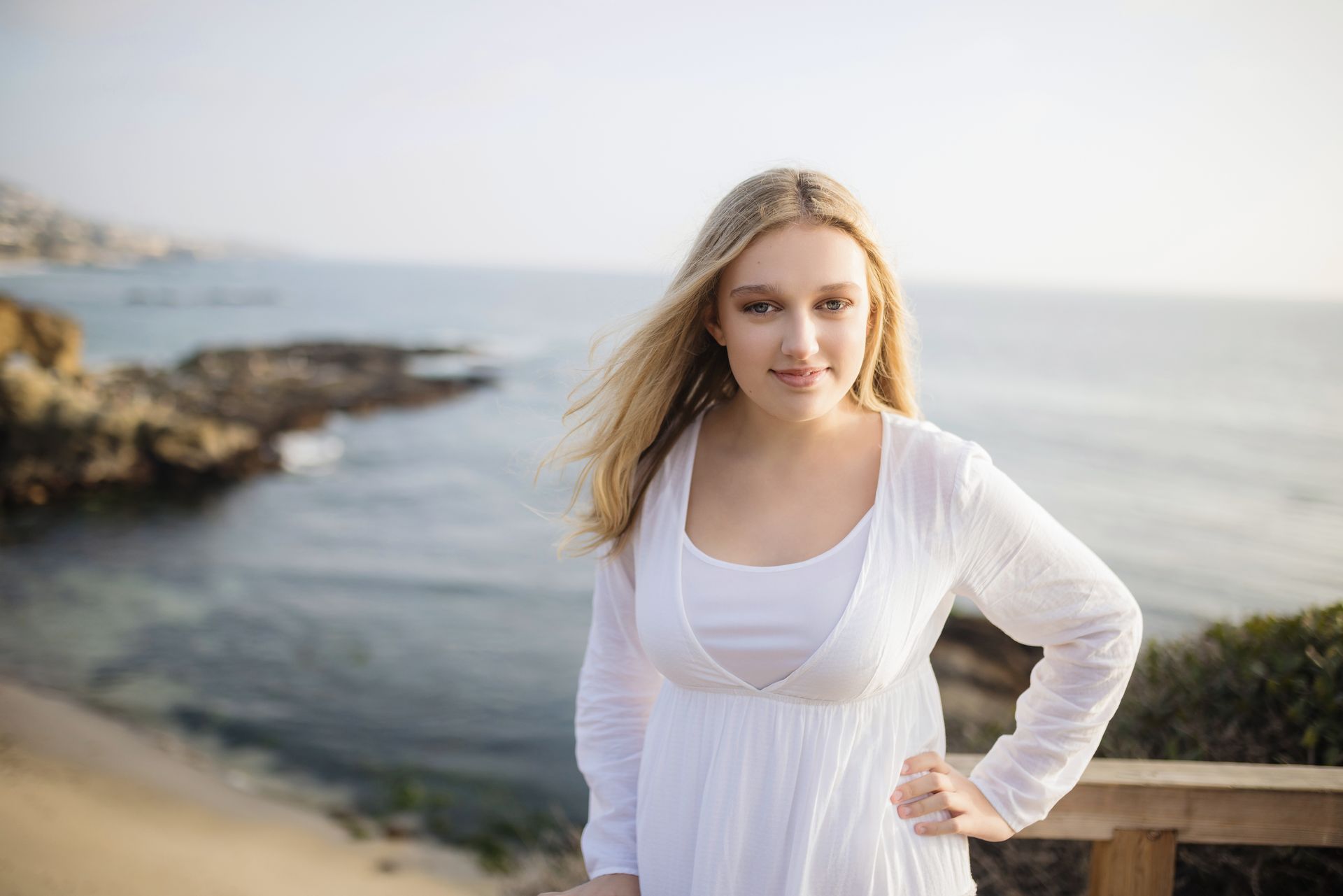 A woman in a white dress is standing on a bridge overlooking the ocean.