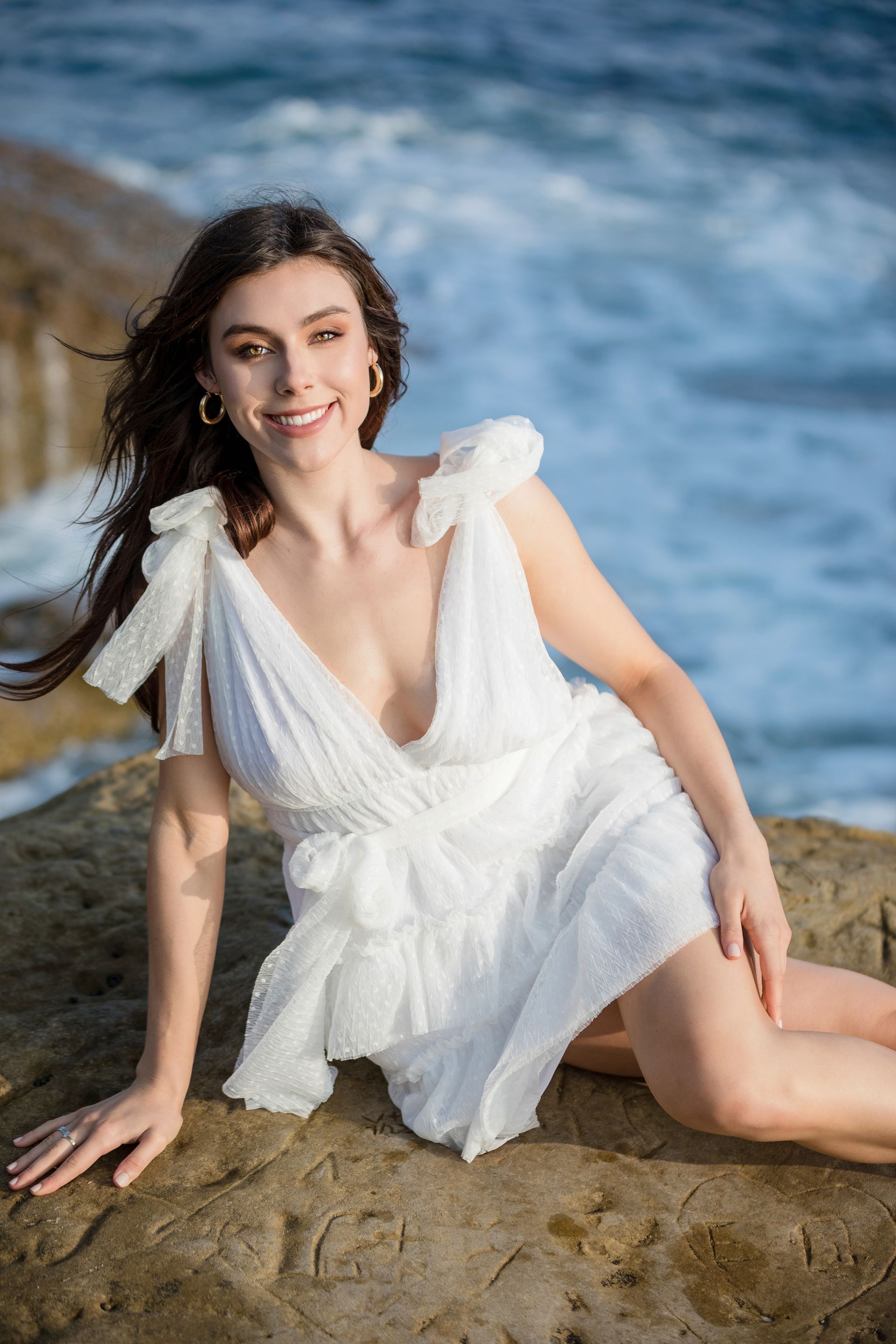 A woman in a white dress is sitting on a rock near the ocean.