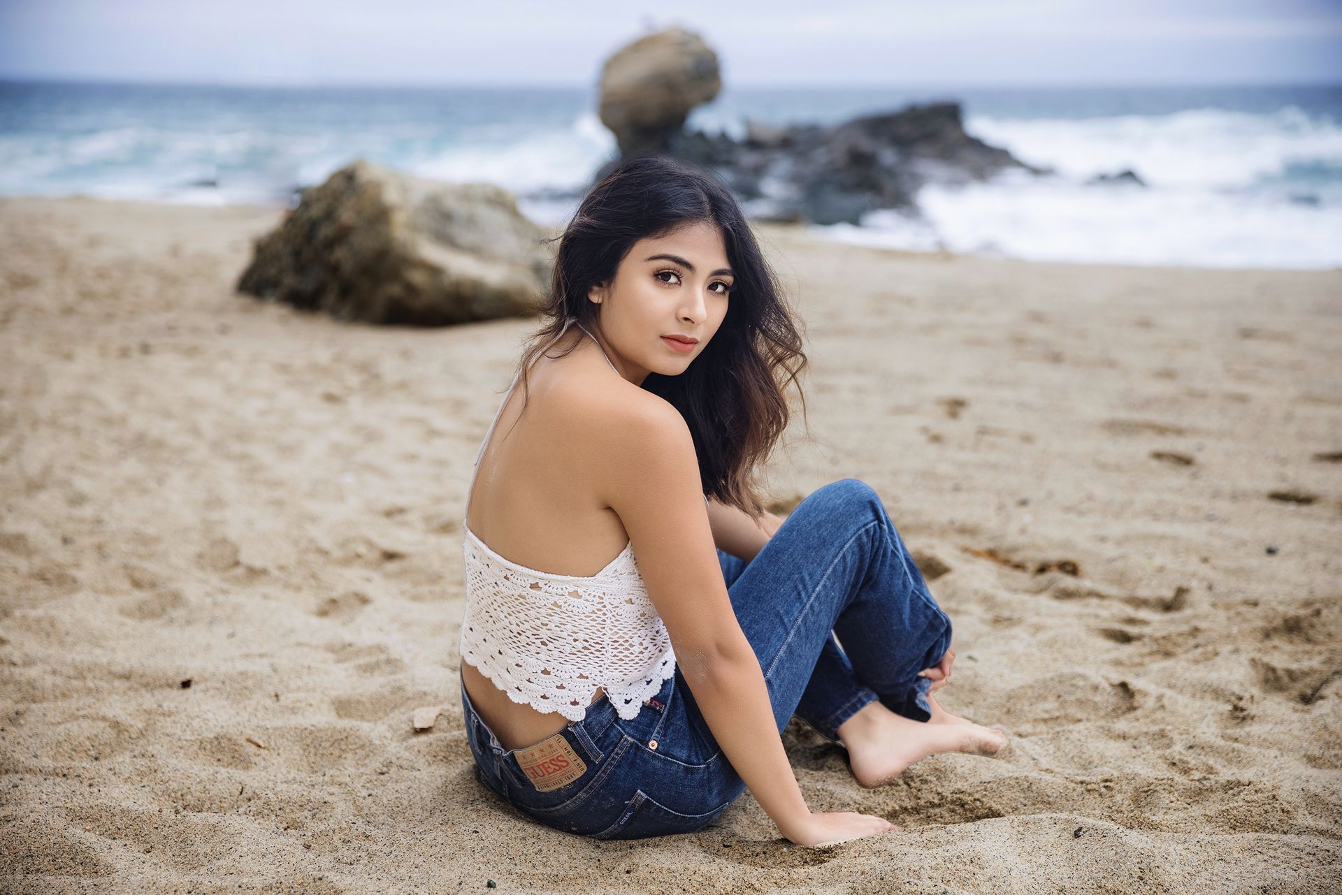 A woman is sitting on the beach wearing jeans and a white top.