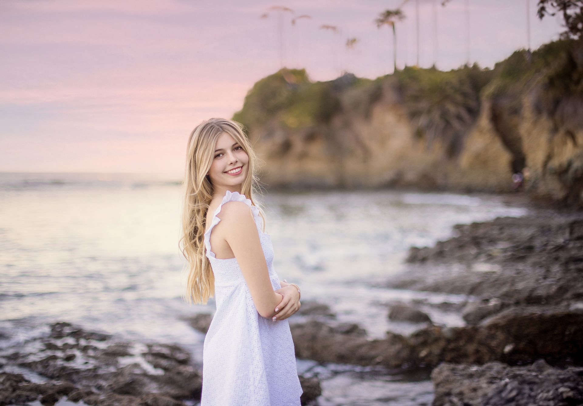 A woman in a white dress is standing on a rocky beach near the ocean.