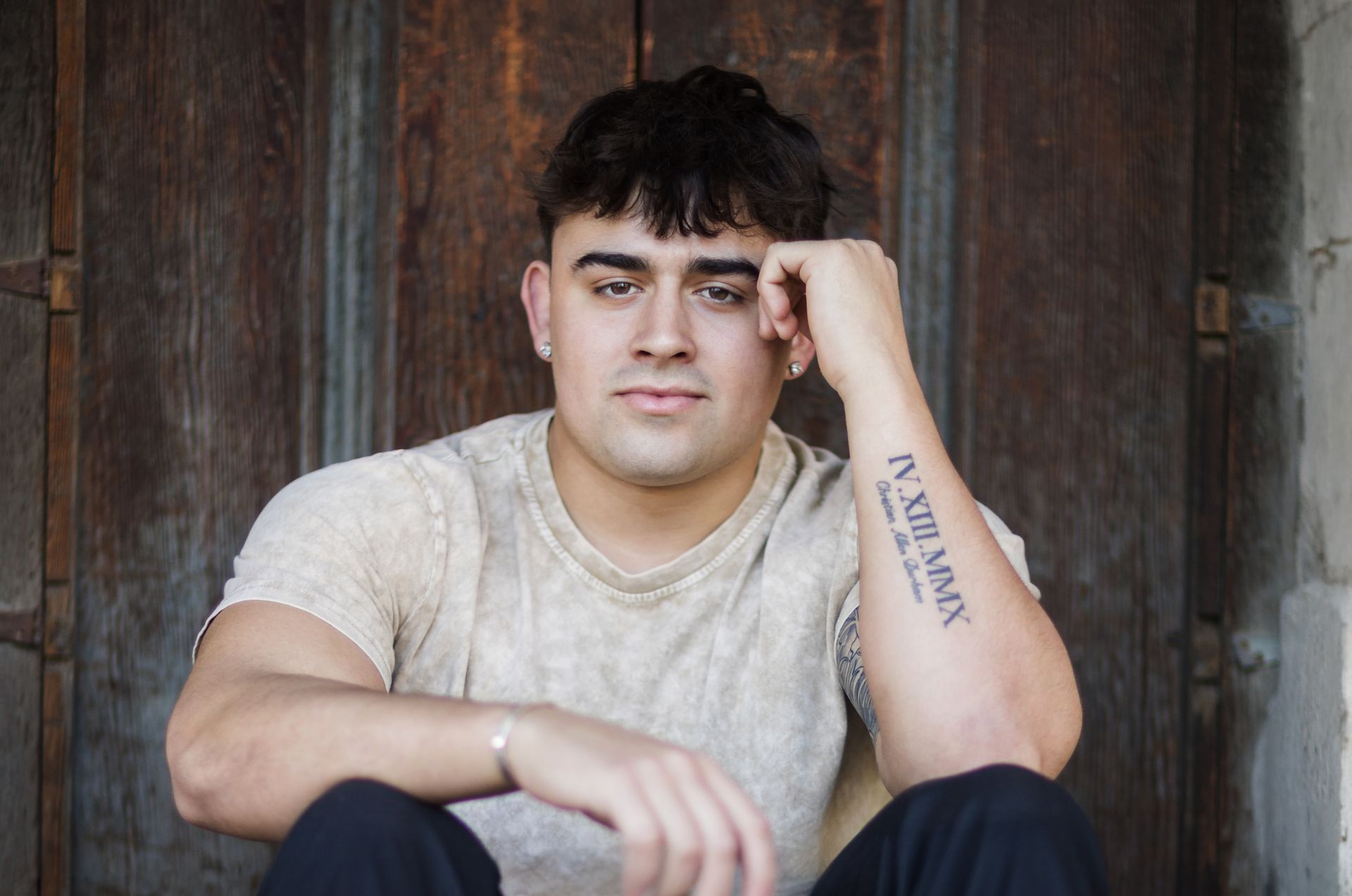 A young man with a tattoo on his arm is sitting in front of a wooden door.