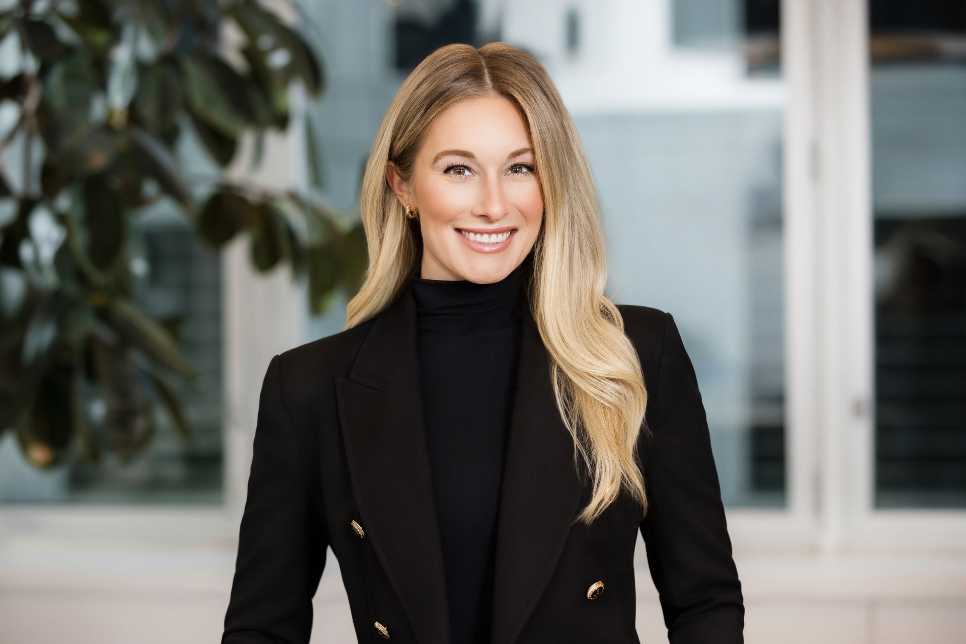 A woman in a black jacket and black turtleneck is smiling in front of a window.