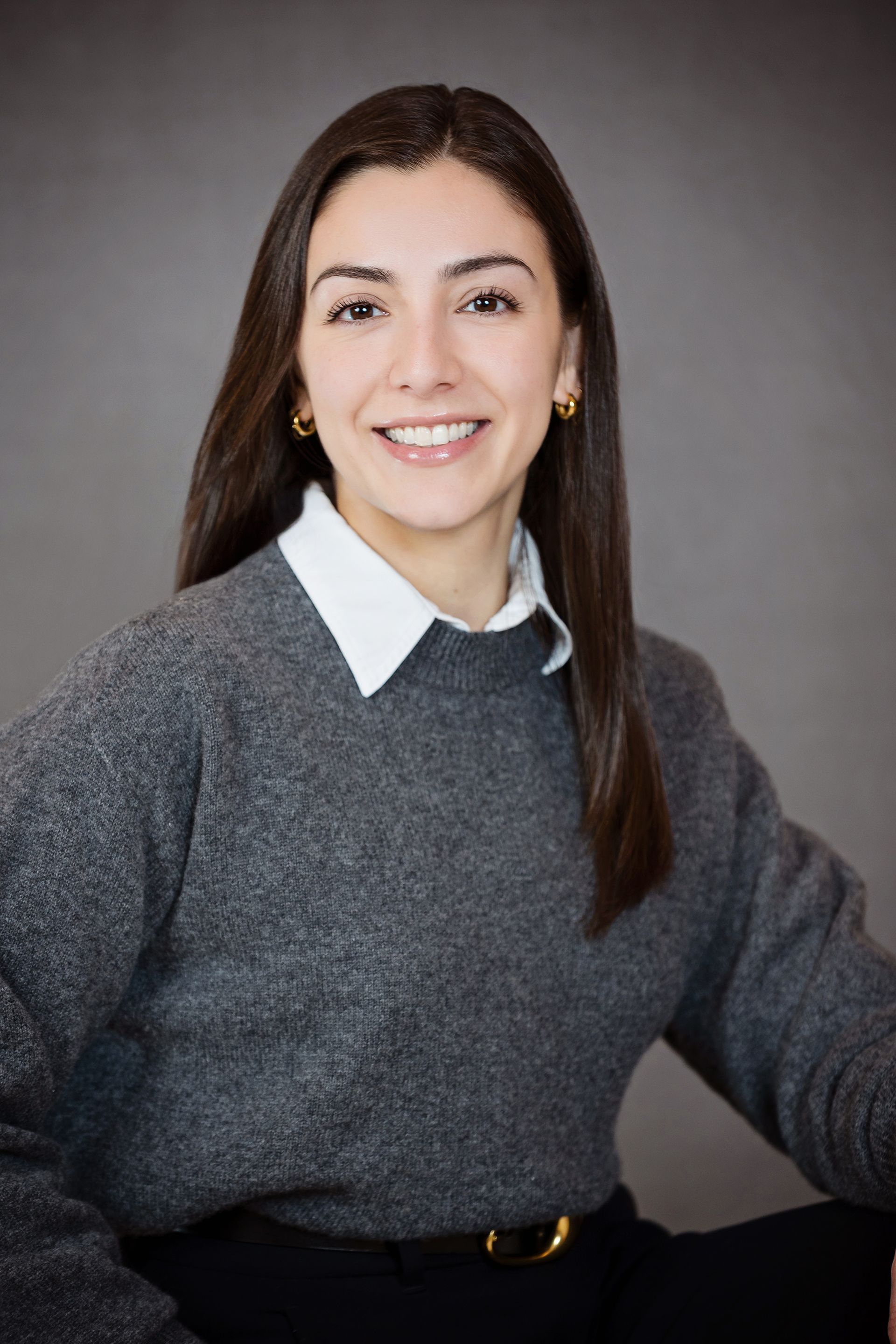 A woman wearing a grey sweater and a white shirt is smiling for the camera.
