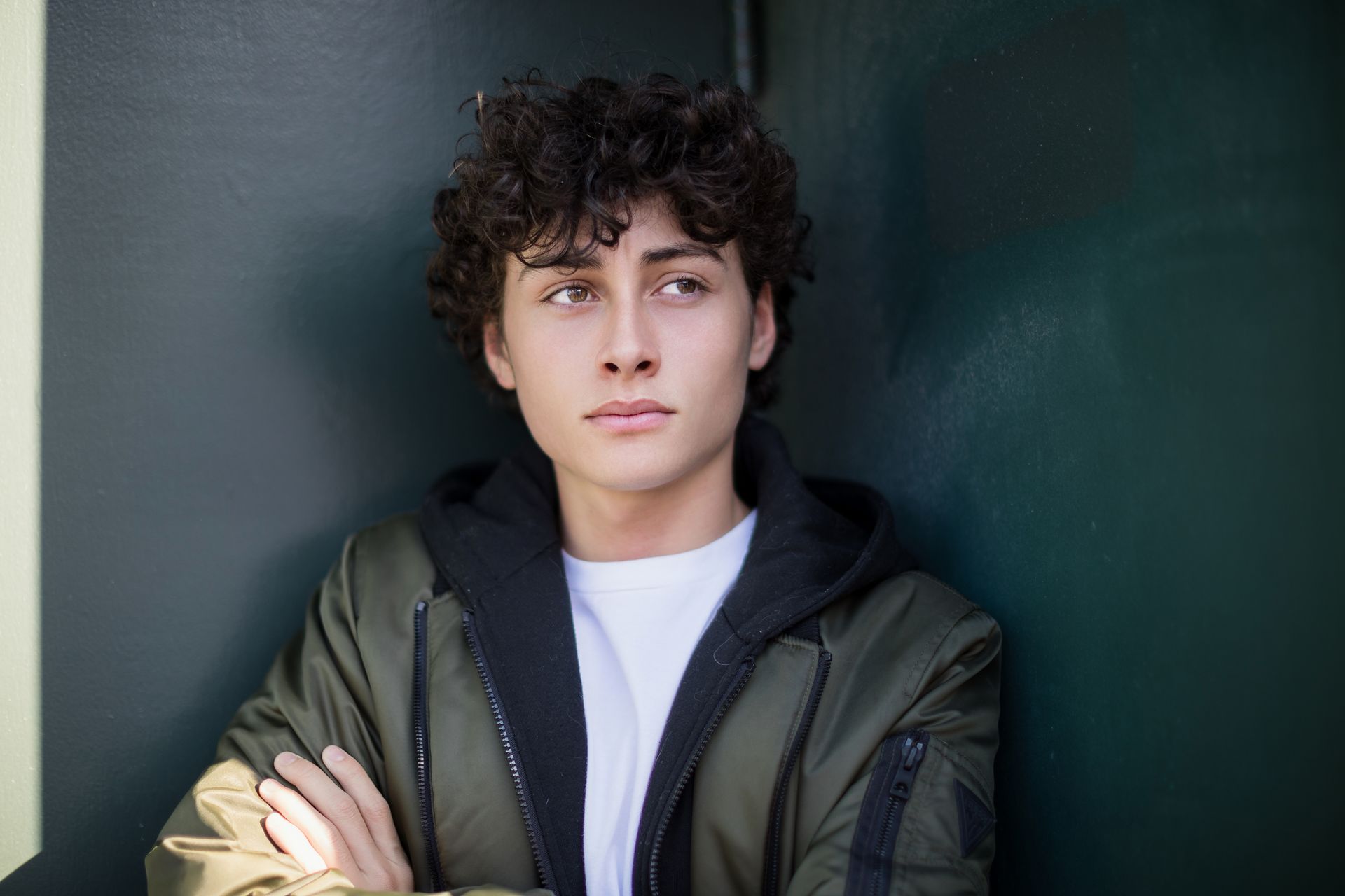 A young man with curly hair is leaning against a wall with his arms crossed.