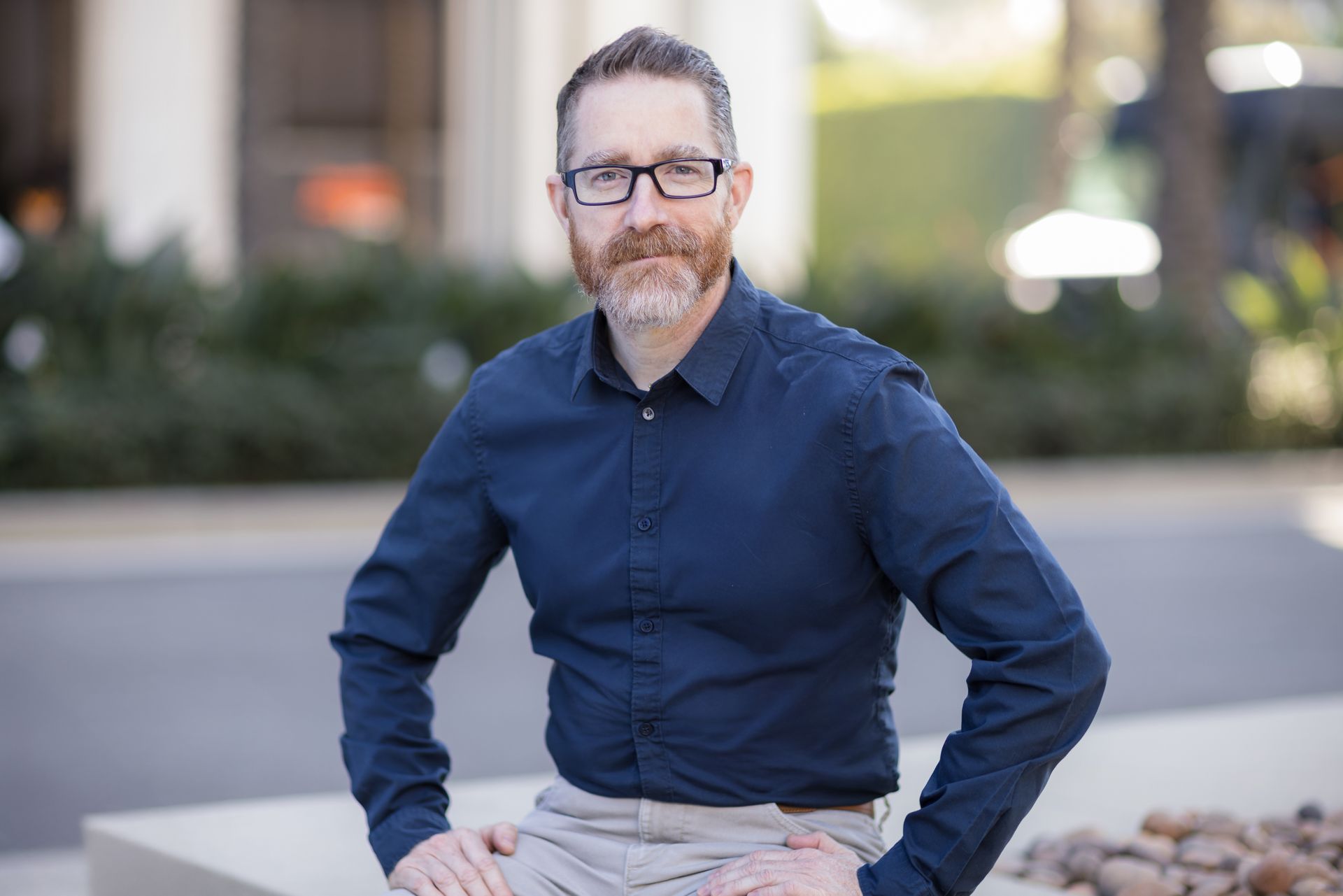 A man wearing glasses and a blue shirt is sitting on a bench with his hands on his hips.