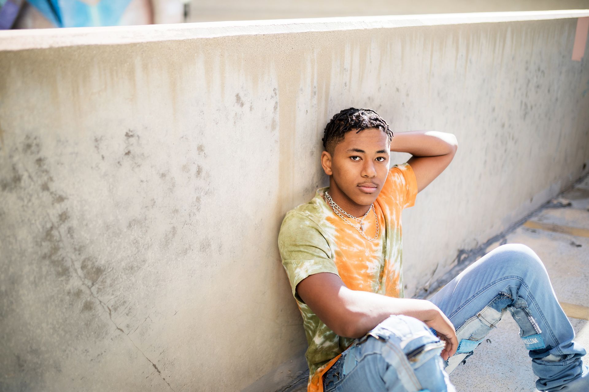 A young man is sitting on the ground leaning against a wall.