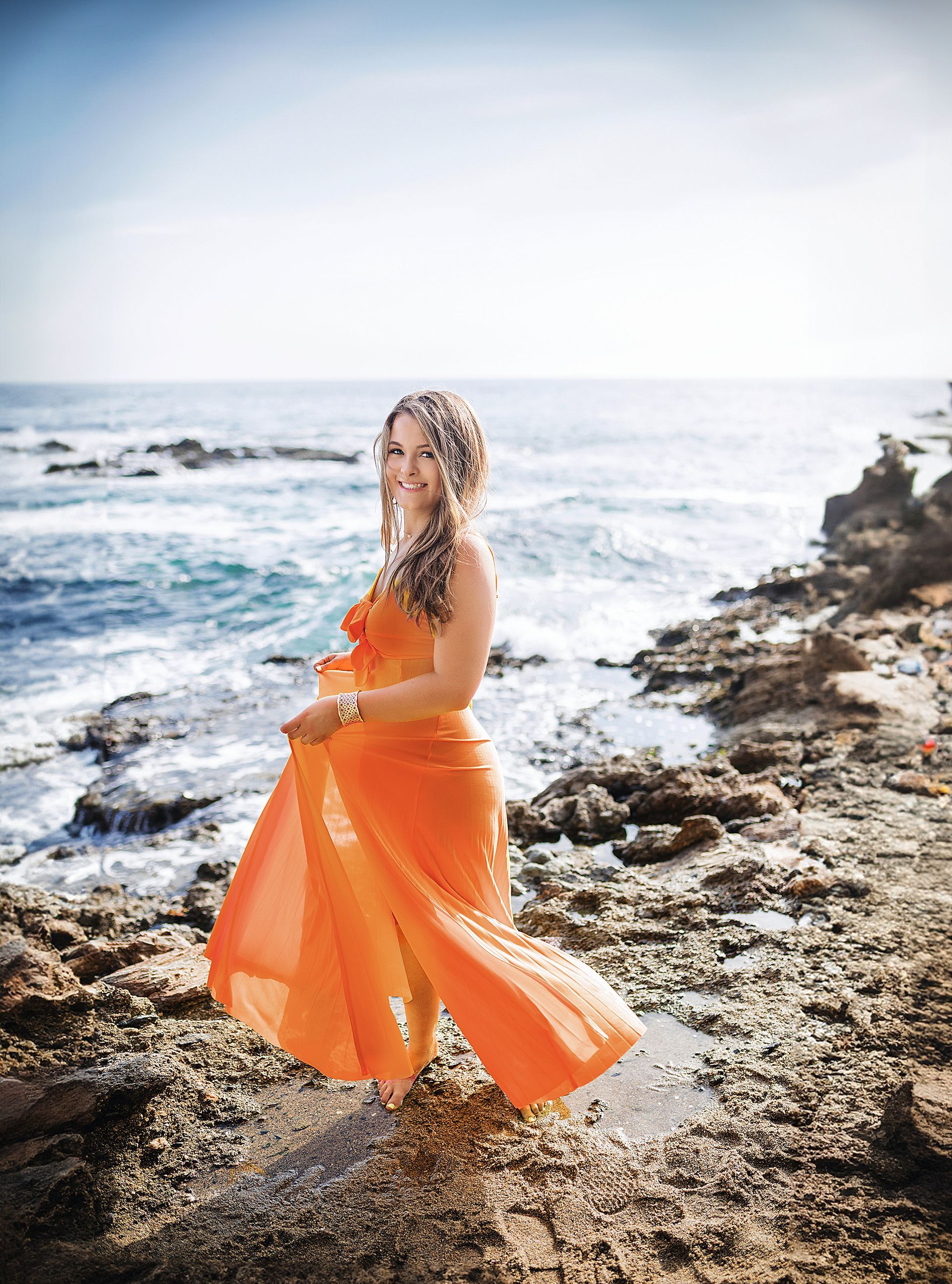 A woman in an orange dress is standing on a rocky beach near the ocean.
