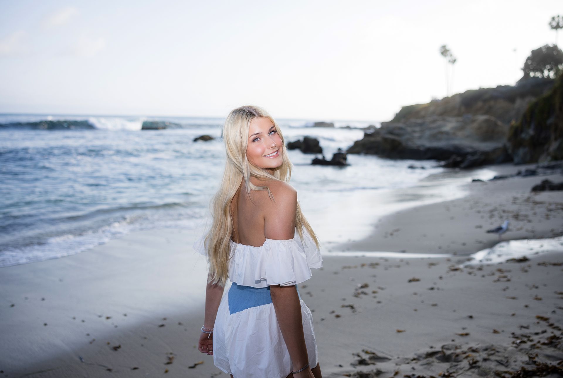 Woman in a white dress standing on a beach at sunset, looking back toward the camera.