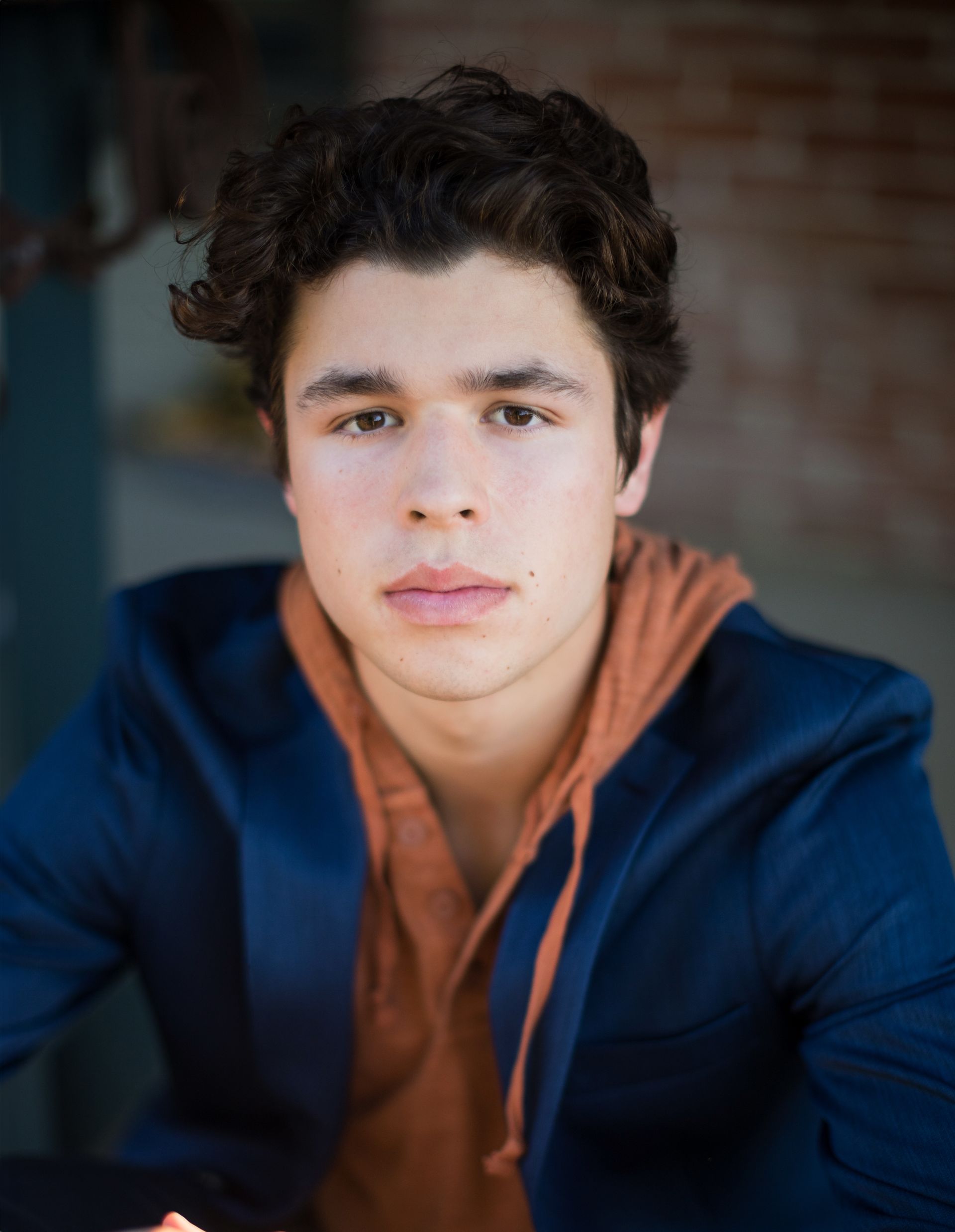 A young man wearing a blue jacket and an orange hoodie is looking at the camera.