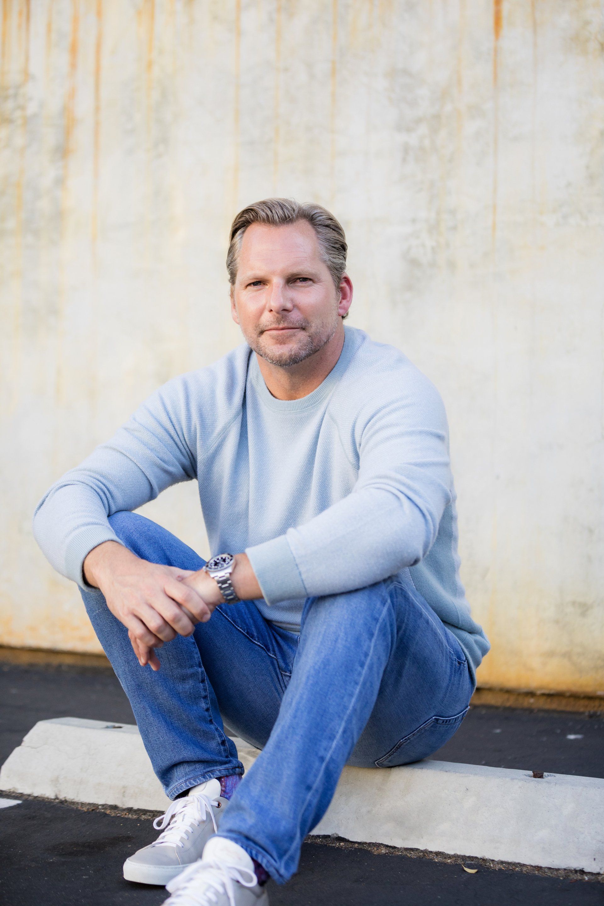 A man is sitting on the curb wearing a blue sweater and jeans.