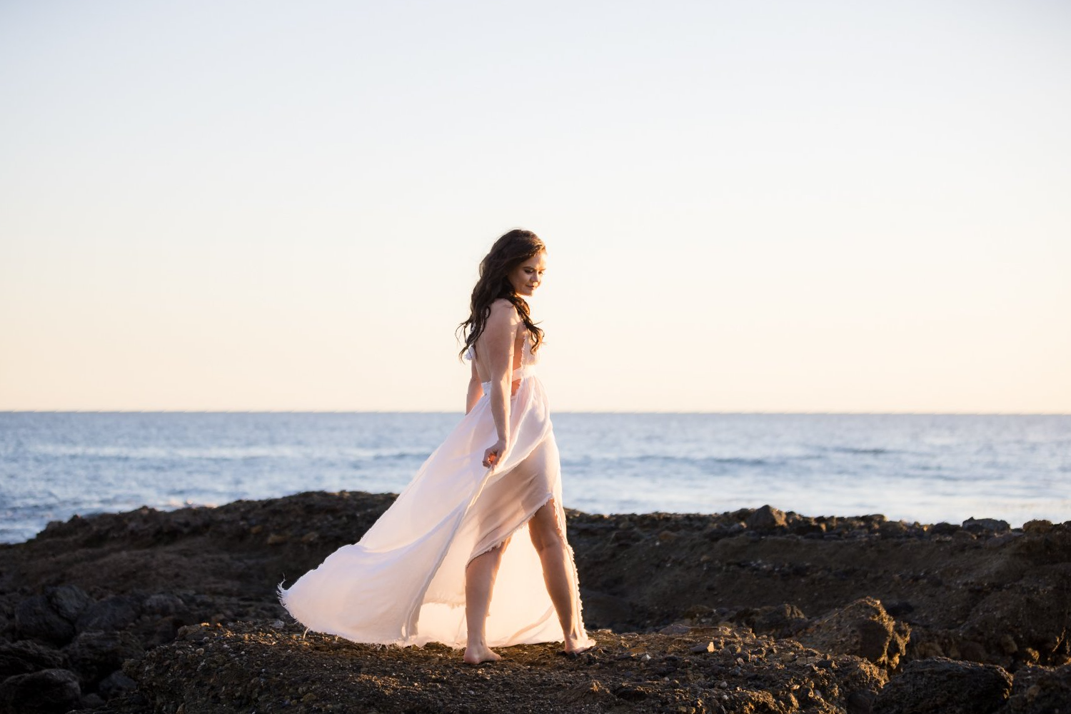 A woman in a long white dress is standing on a rocky beach near the ocean.