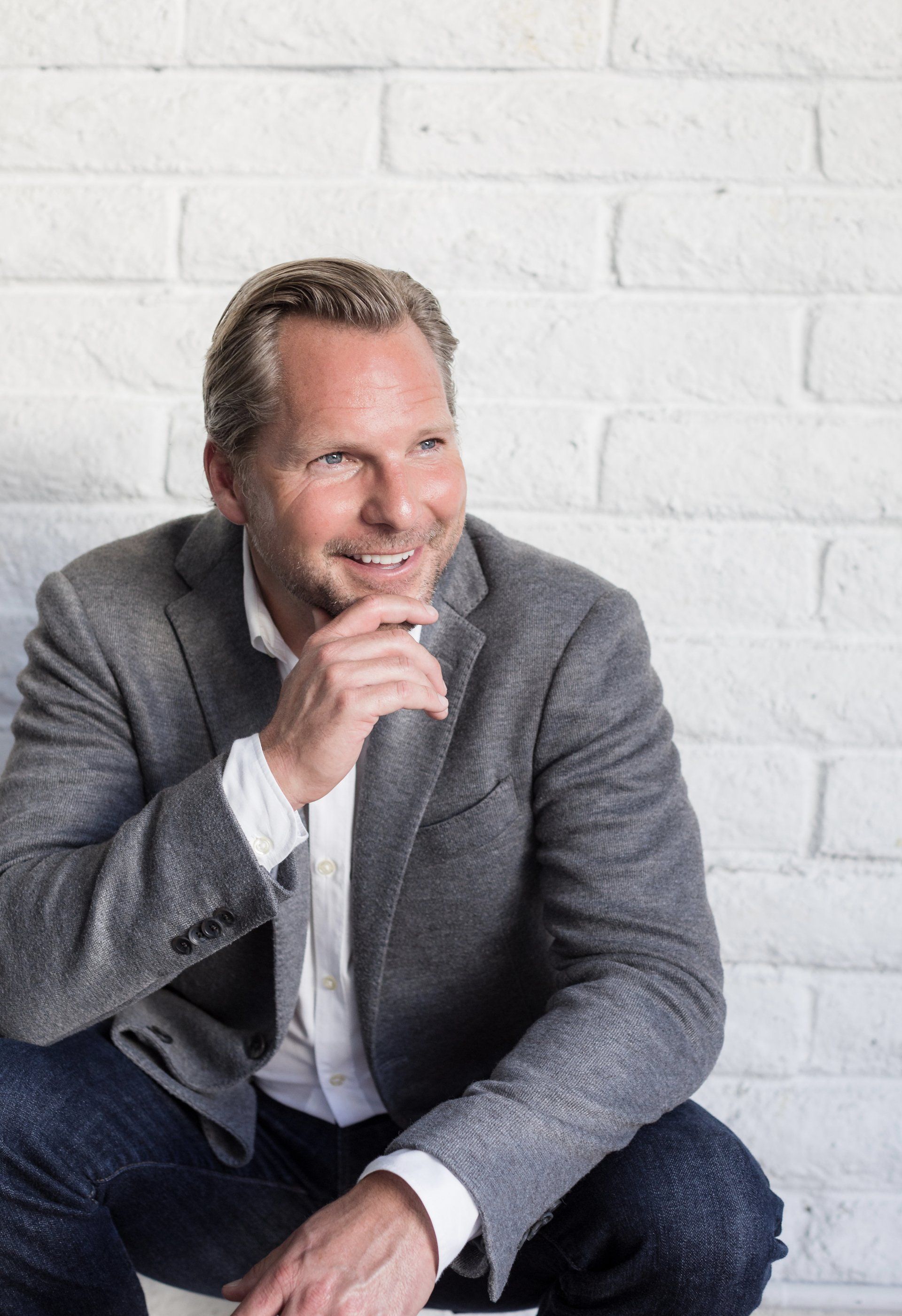 Relaxed lifestyle portrait of a man in a grey blazer against a white brick wall
