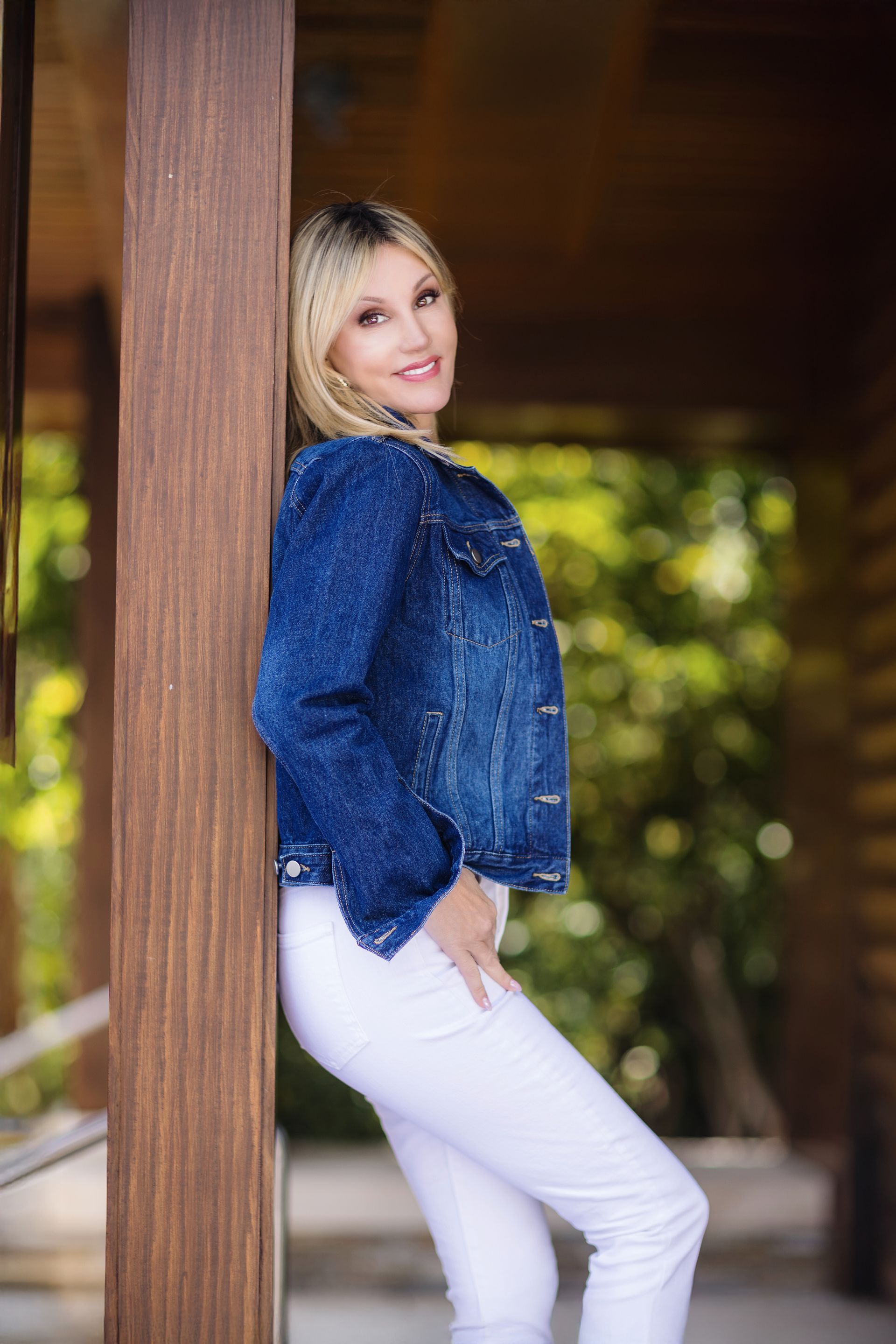 A woman in a denim jacket and white jeans is leaning against a wooden post.