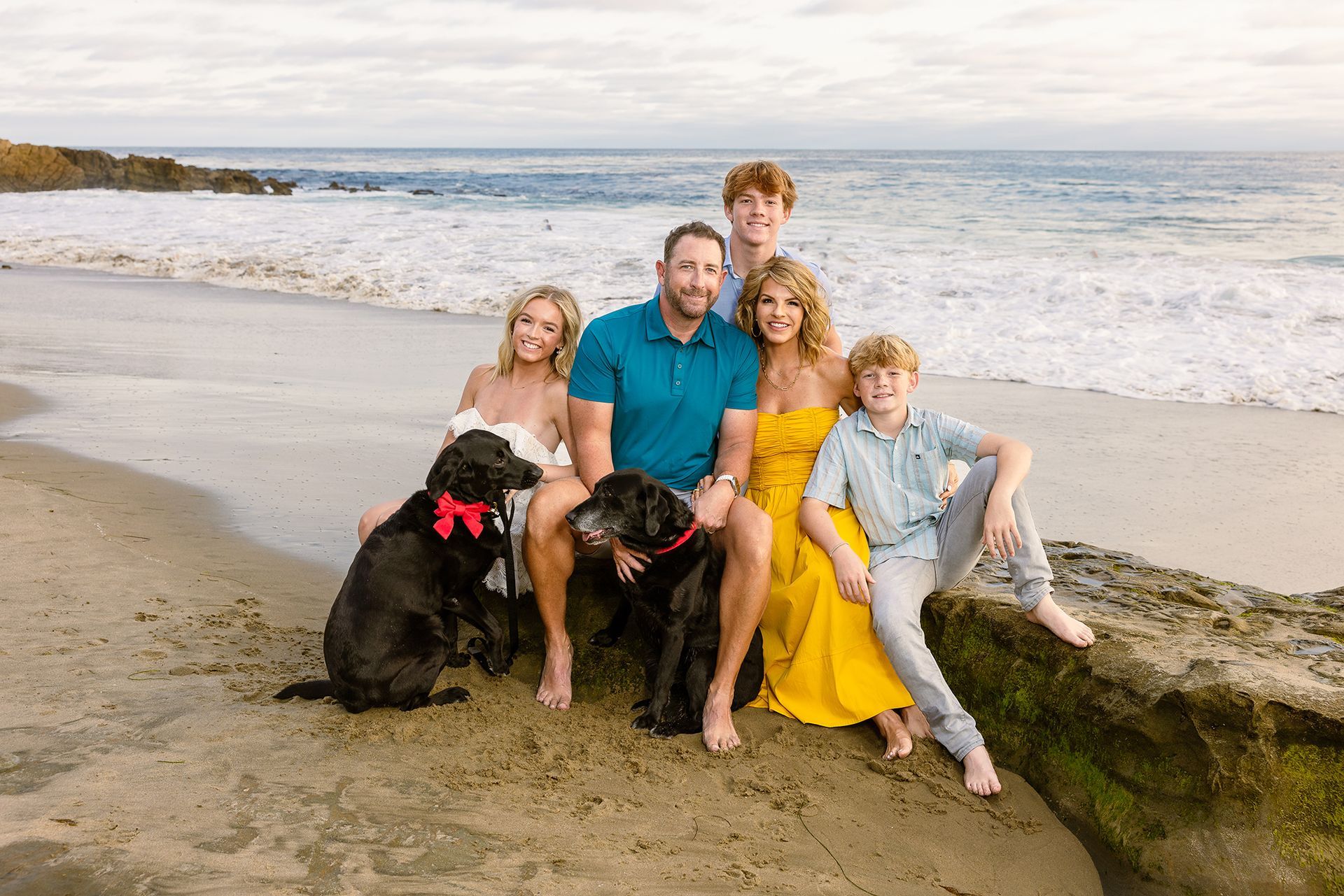 Family of five and two black dogs sitting on a beach near the ocean at sunset