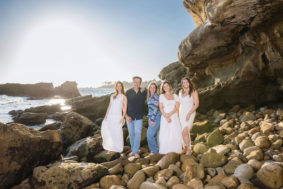 Family standing on rocky beach in Laguna Beach during golden hour, wearing coordinated white and blue outfits