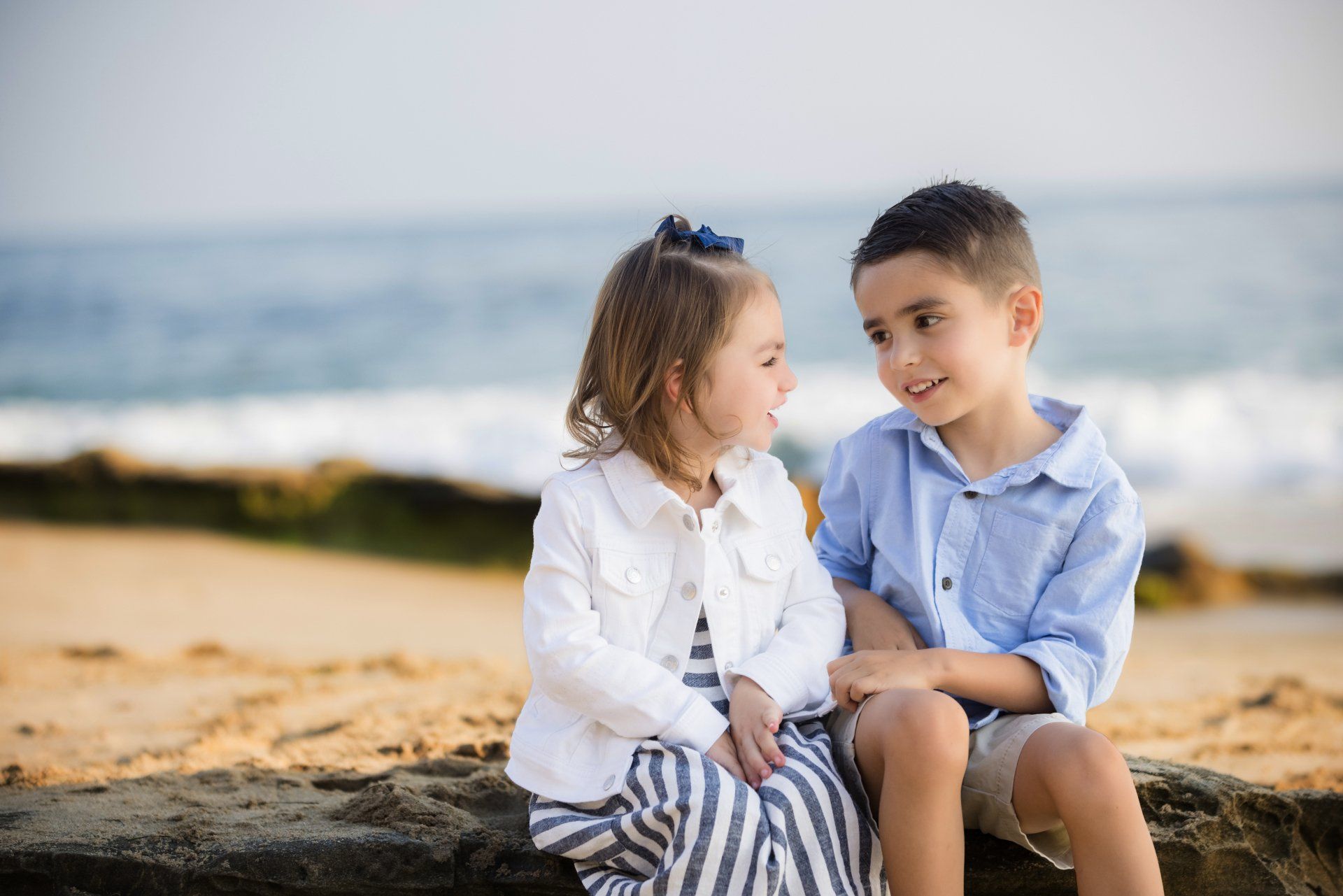 A boy and a girl are sitting on a rock on the beach looking at each other.