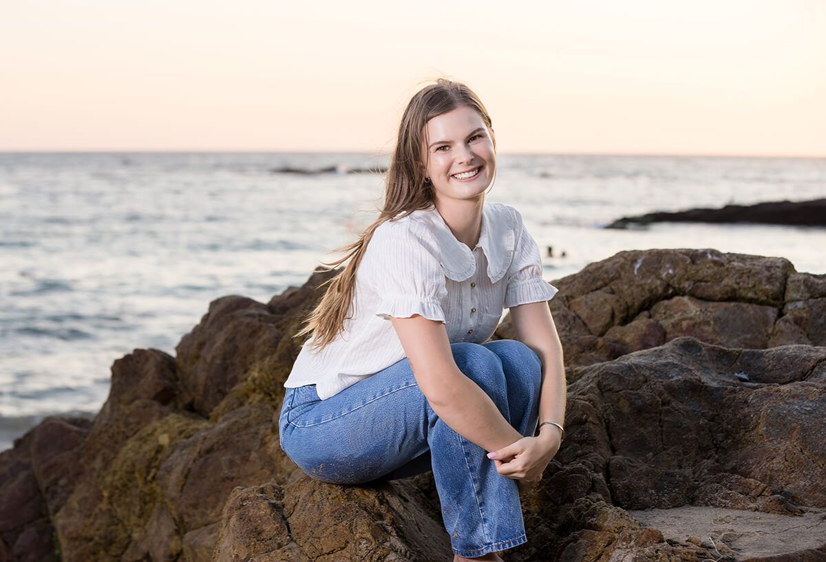 young woman sitting on coastal rocks in Laguna Beach at Golden hour for beach portraits, dressed in a white blouse and jeans