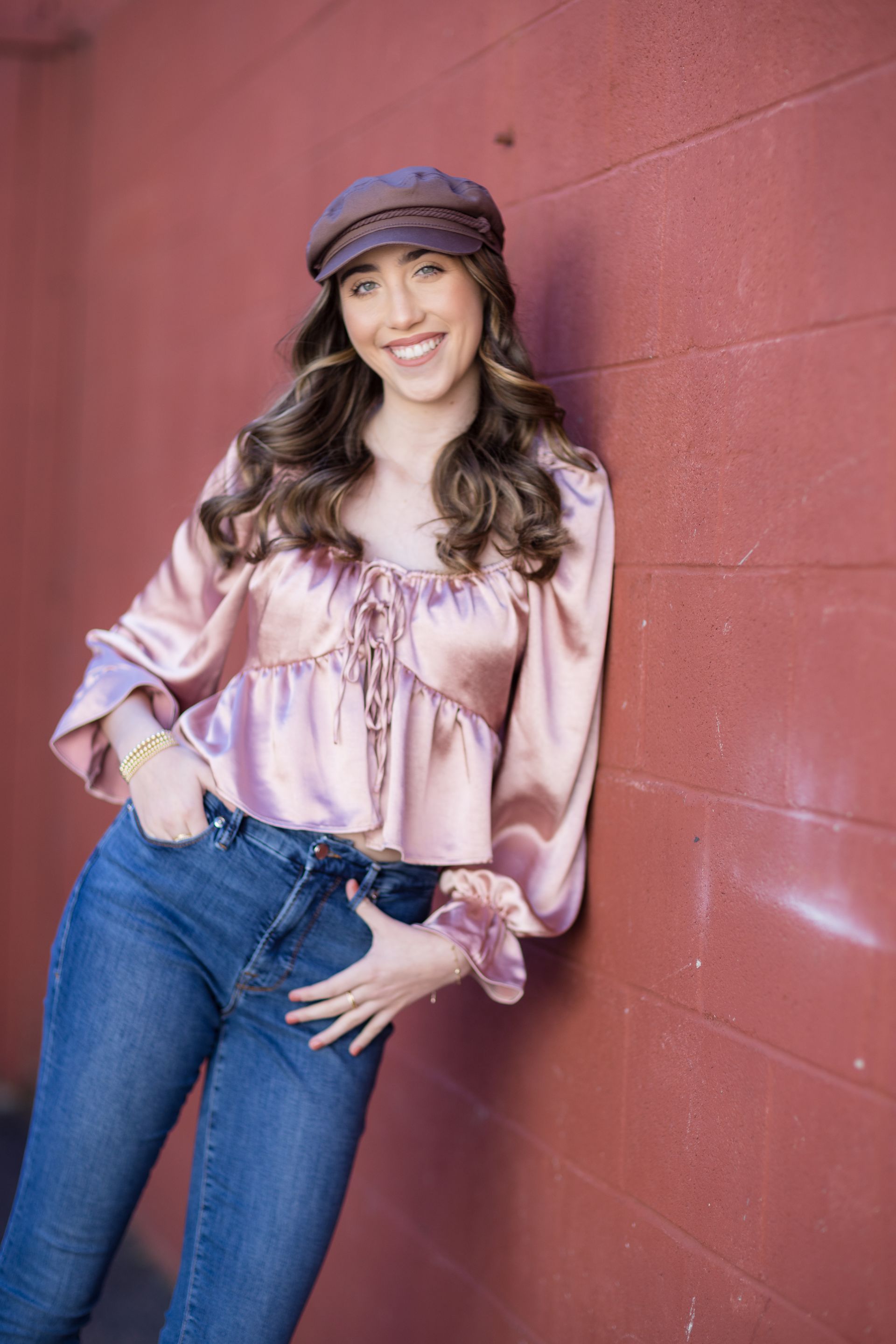 A woman wearing a pink satin top and blue jeans is leaning against a red wall.