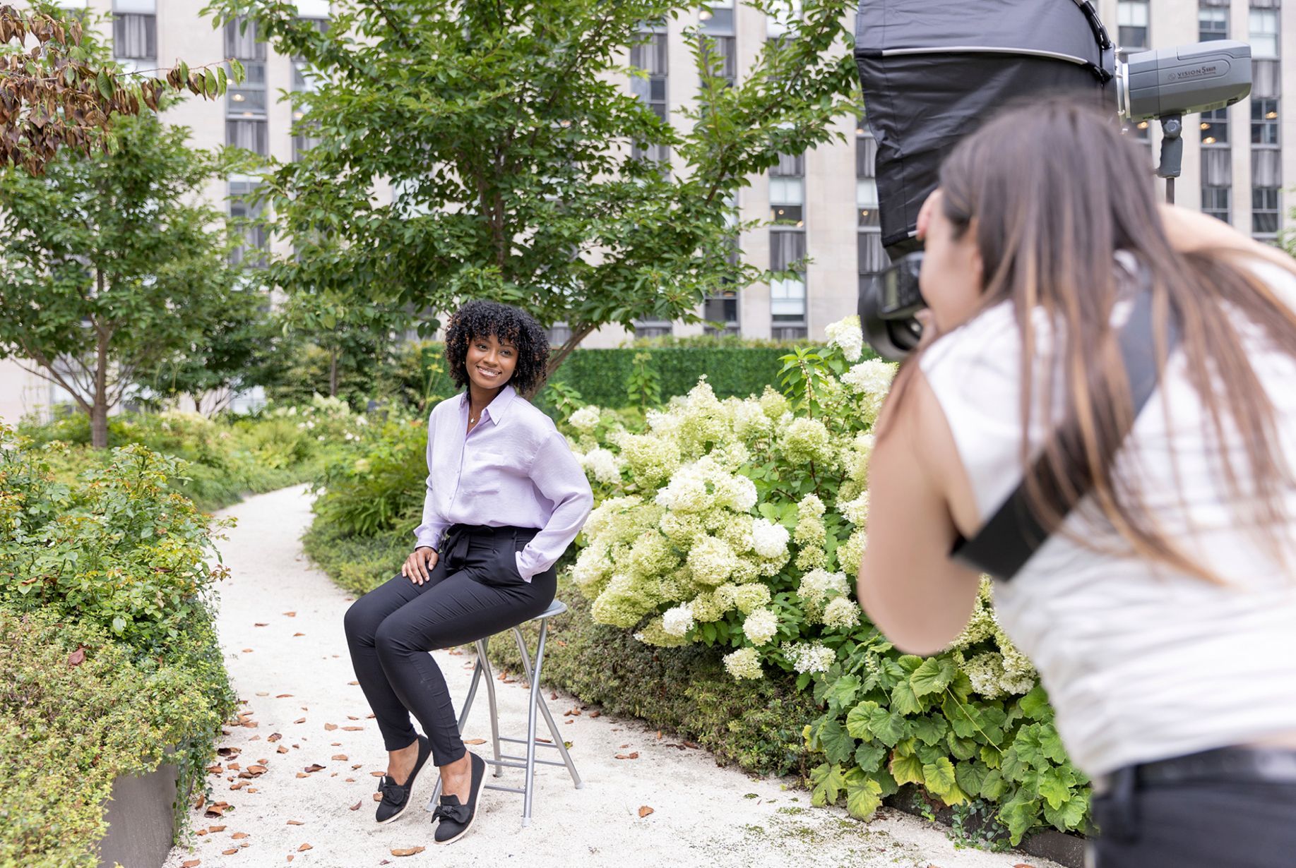 A woman is taking a picture of a woman sitting on a stool in a park.