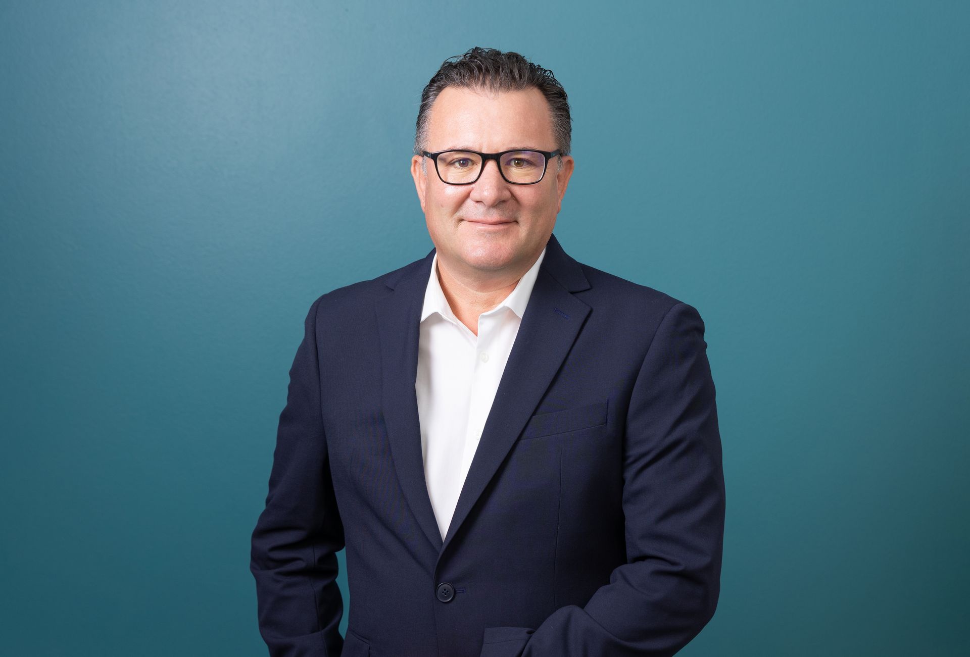 A man in a suit and glasses is standing in front of a blue wall.