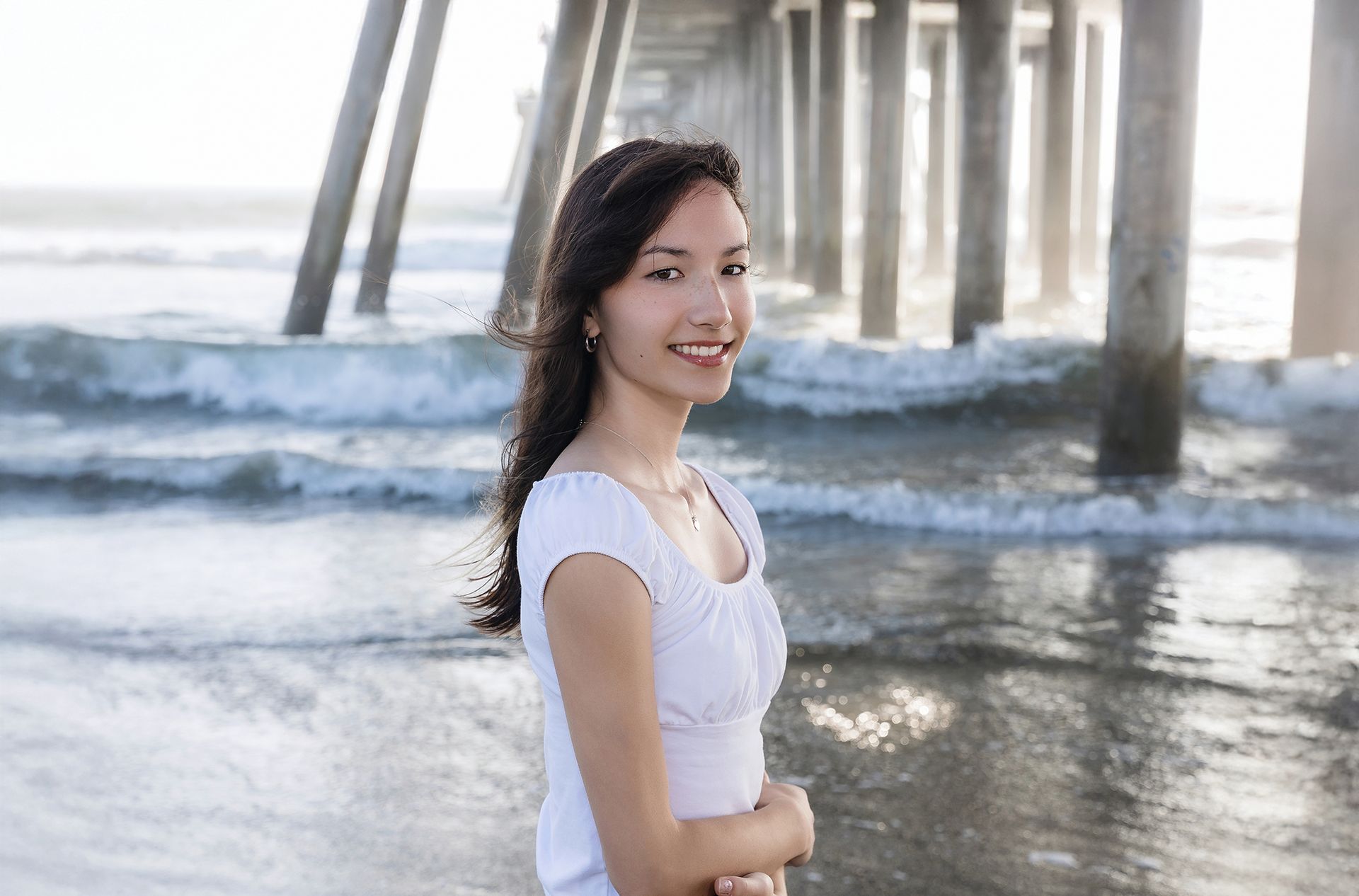 Senior girl in white under the Huntington Beach Pier with waves behind her