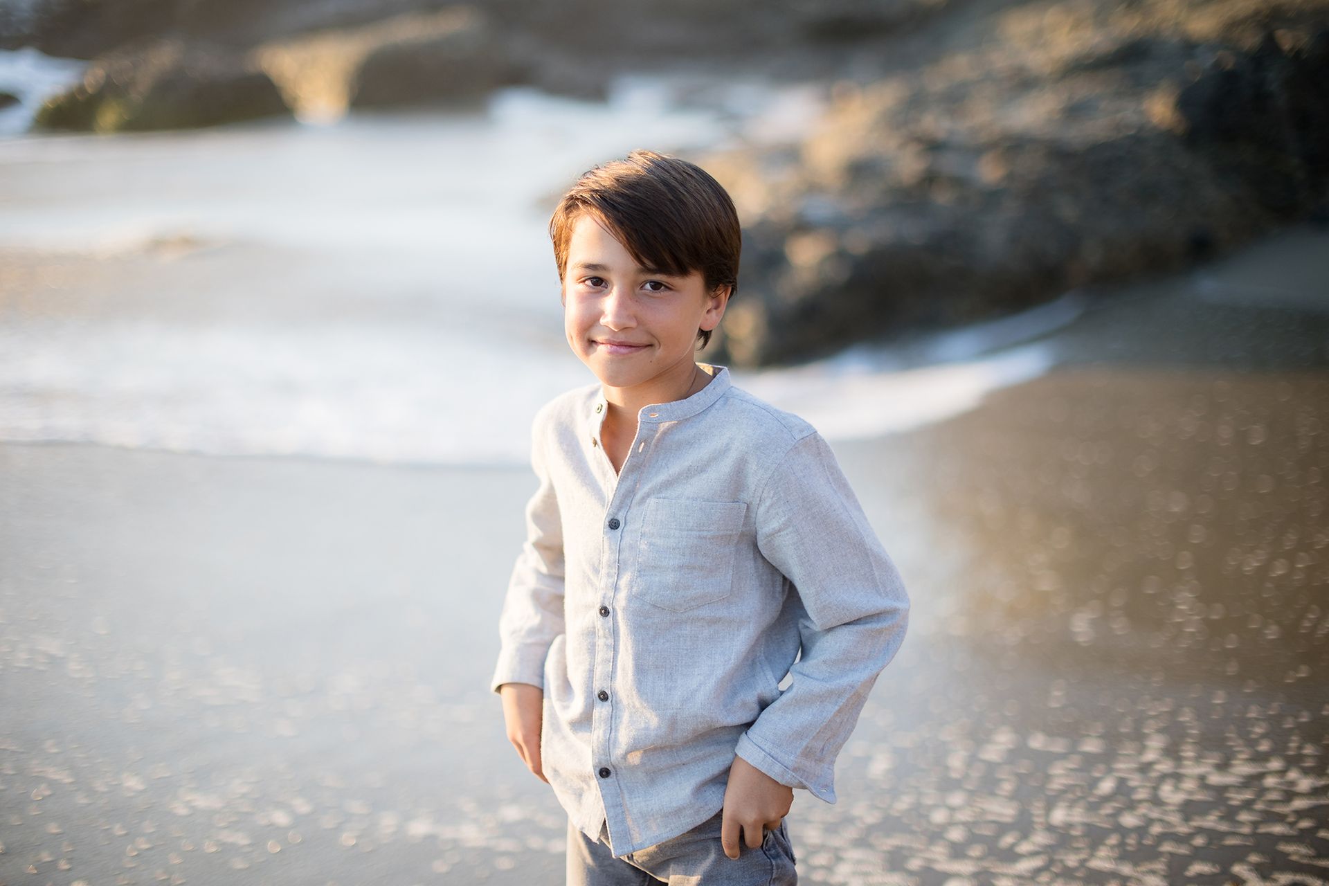 oung boy in soft gray linen shirt posing on the sand during a golden hour session by Orange County family photographer in Laguna Beach.