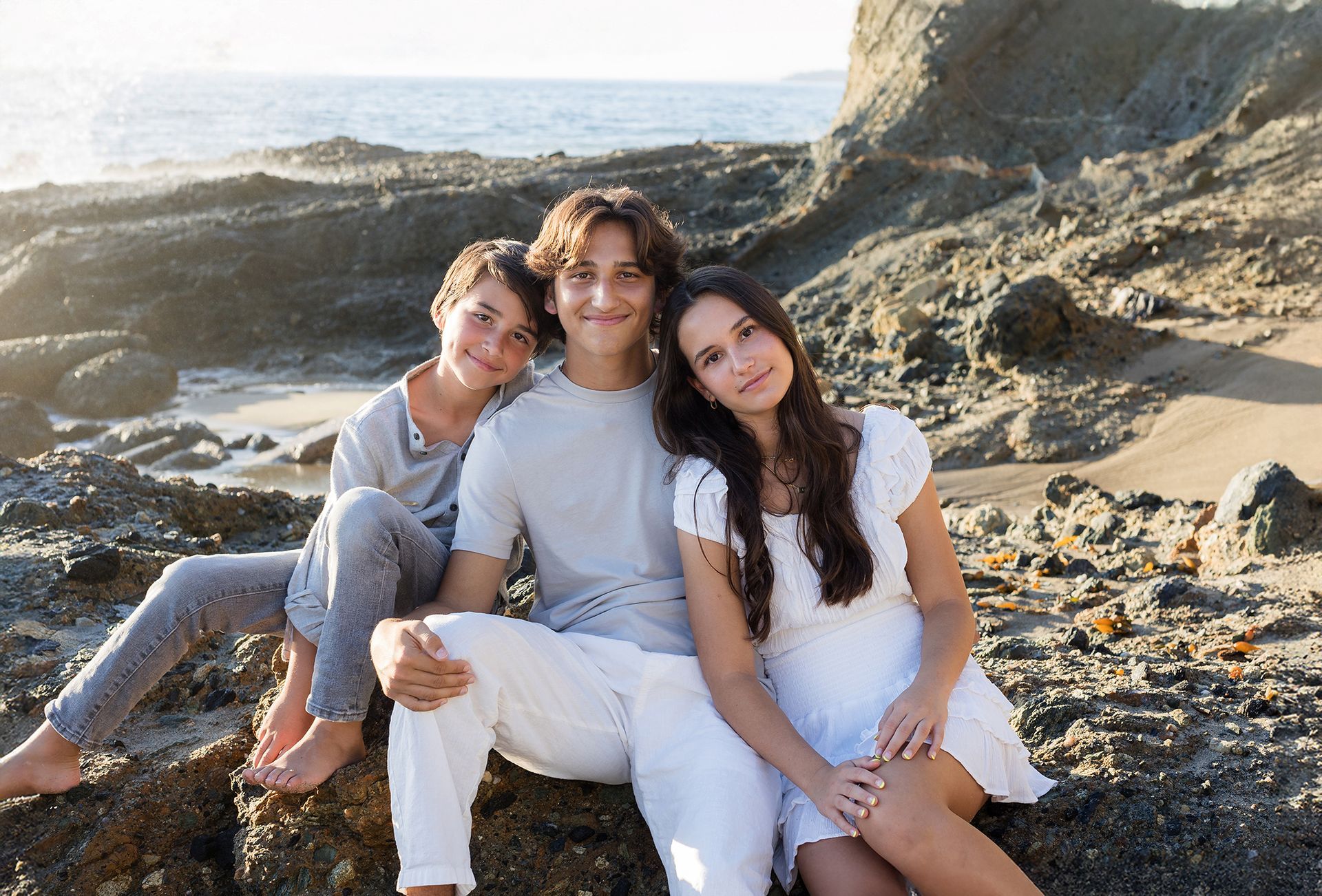 Three people sitting on rocks at a sunny beach, with ocean waves and cliffs in the background.