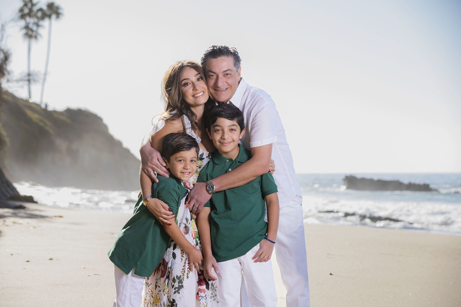 Family of four hugging on a beach, smiling in white and green outfits.