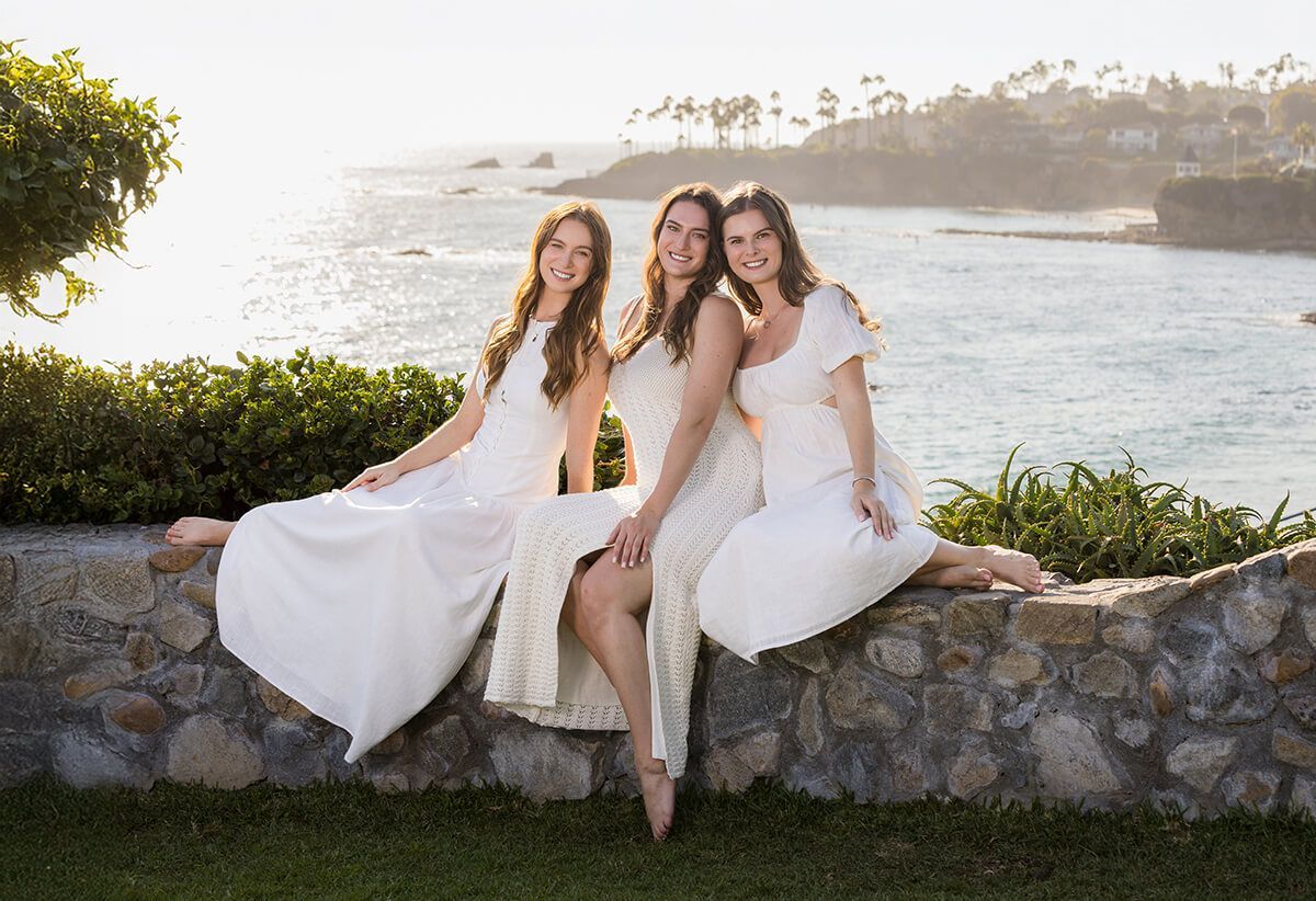 Three sisters in white dresses on the bluff-top stone wall above the Laguna Beach coastline at sunset