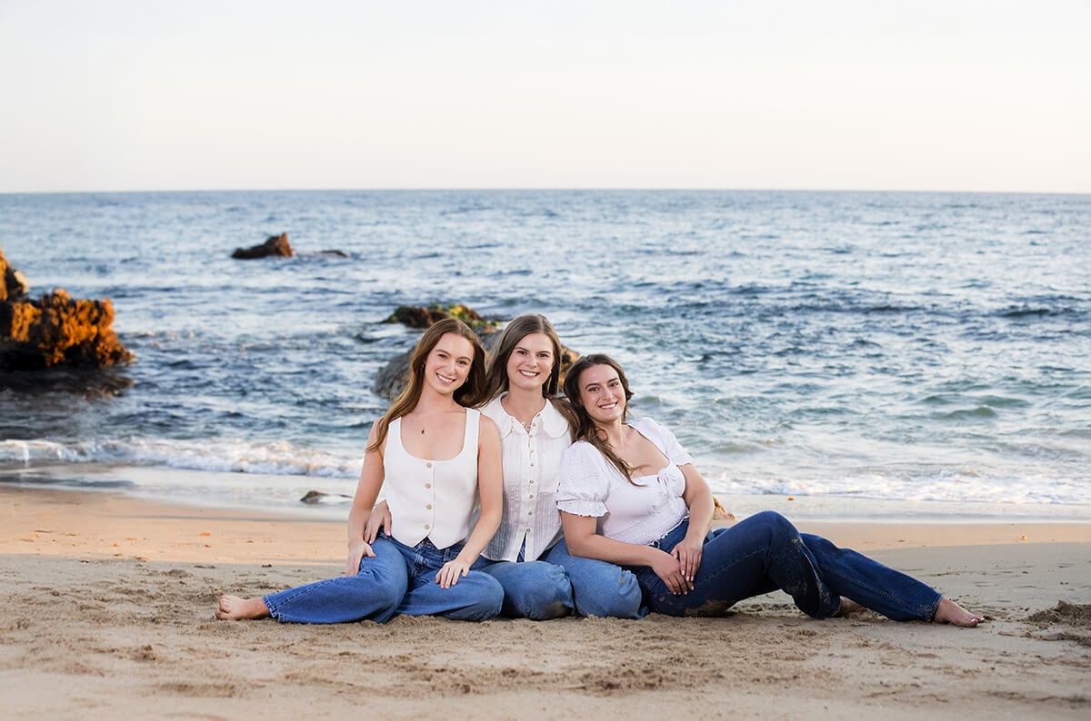 Three sisters sitting on the sand in coordinated white tops and jeans during a sibling beach portrait session in Laguna Beach