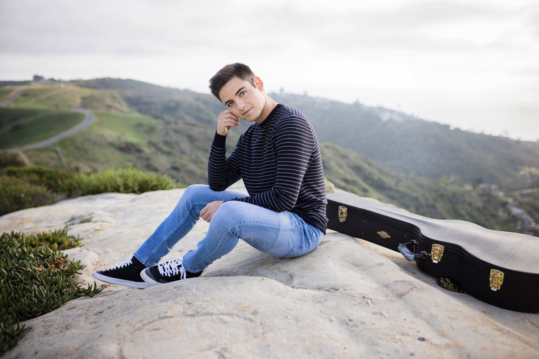 A young man is sitting on a rock with a guitar case.