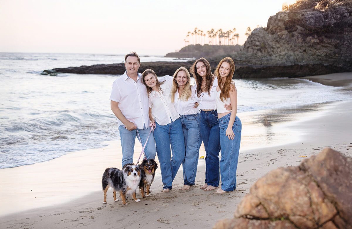 Lifestyle family portrait at sunset on the beach in Laguna Beach with two dogs, featuring parents and three daughters in white tops and denim