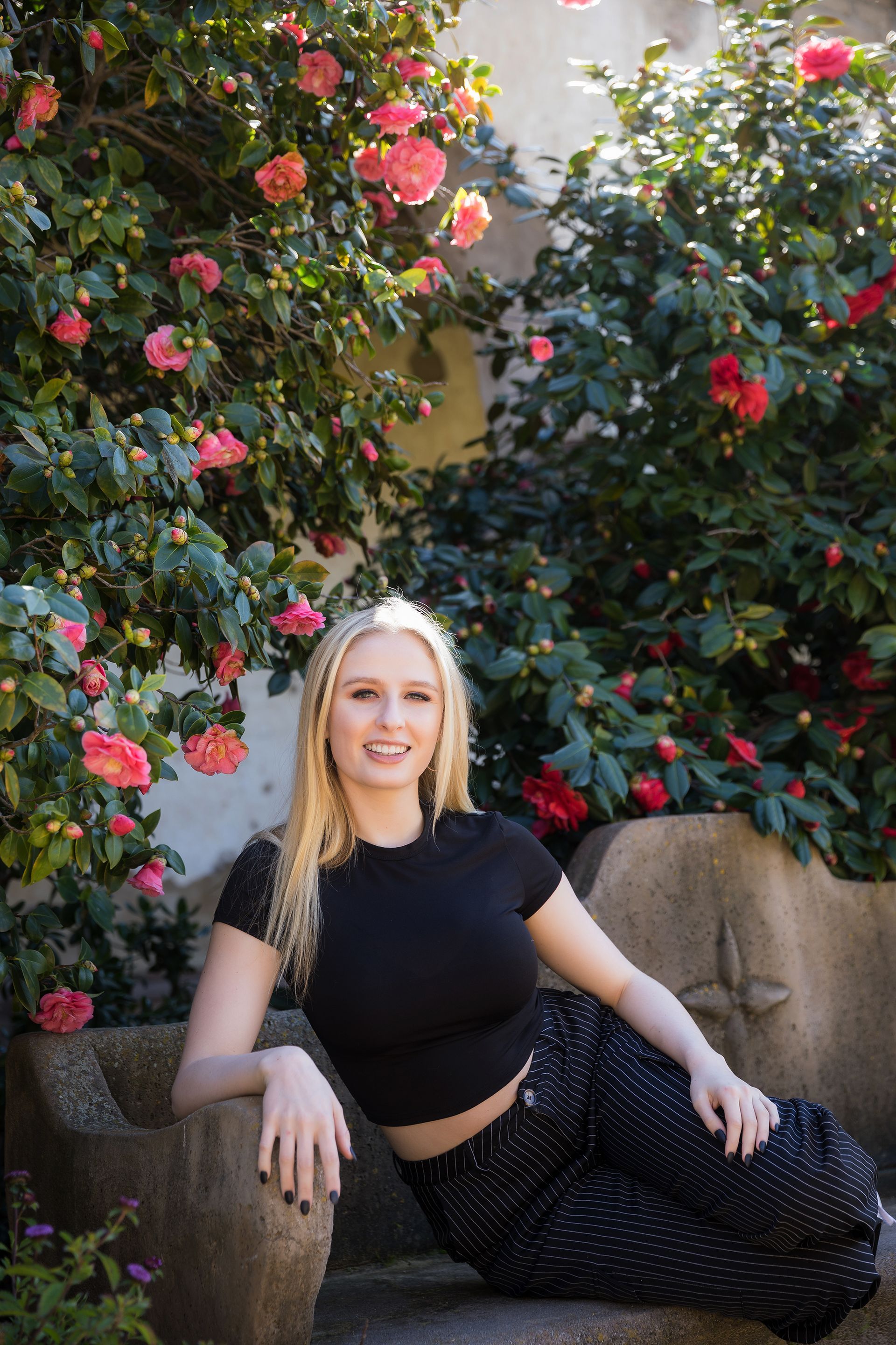 A woman is sitting on a bench in front of a bush of flowers.