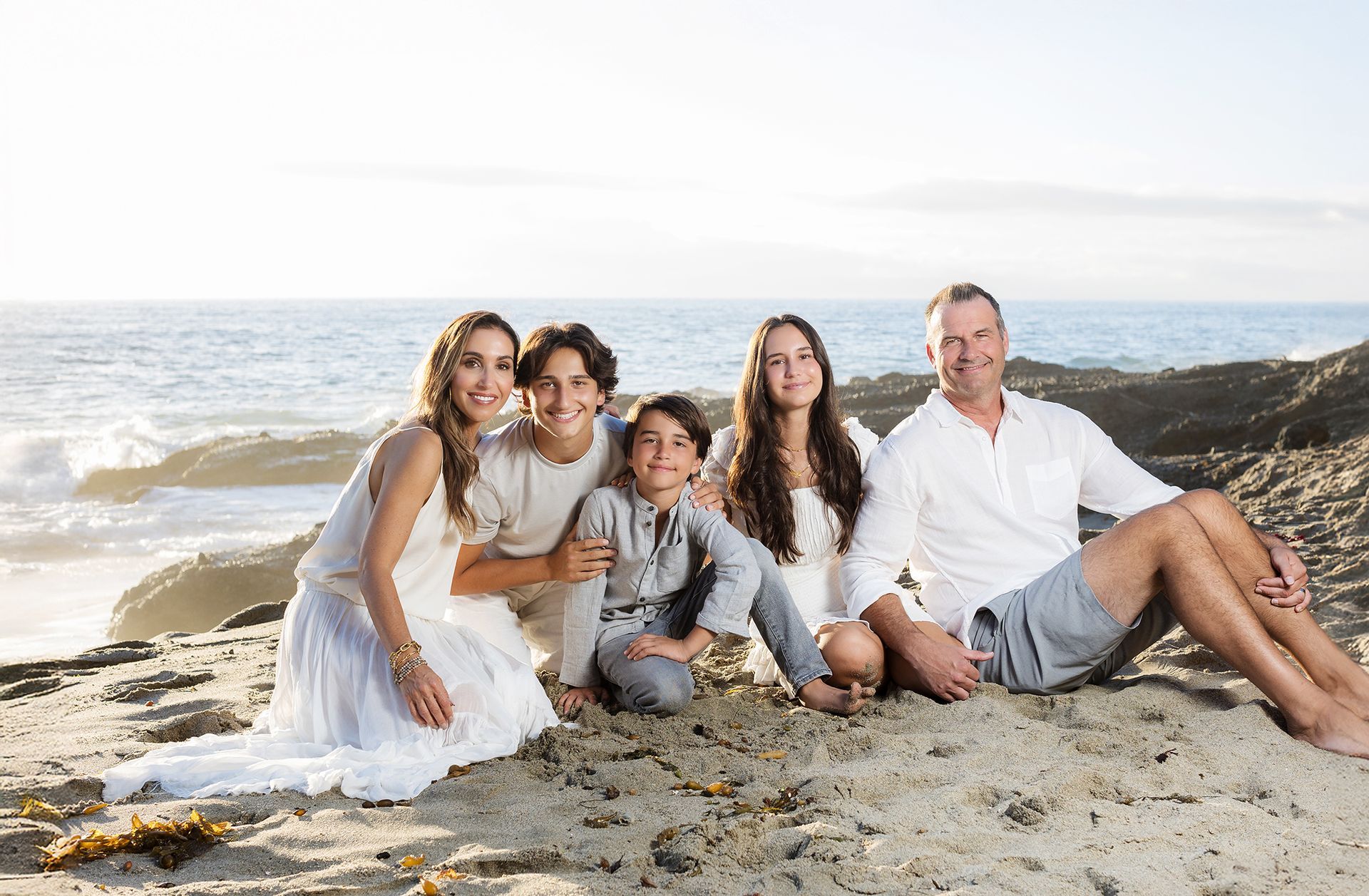Full family seated together on rocky shoreline at The Montage Laguna Beach for high-end family portraits by an Orange County photographer.