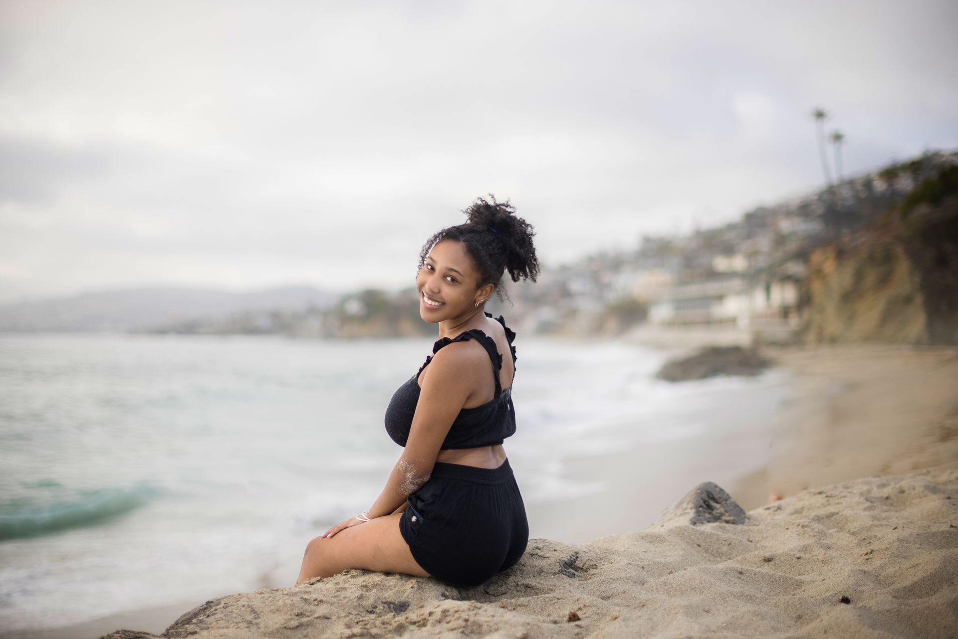A woman is sitting on a rock on the beach looking at the ocean.