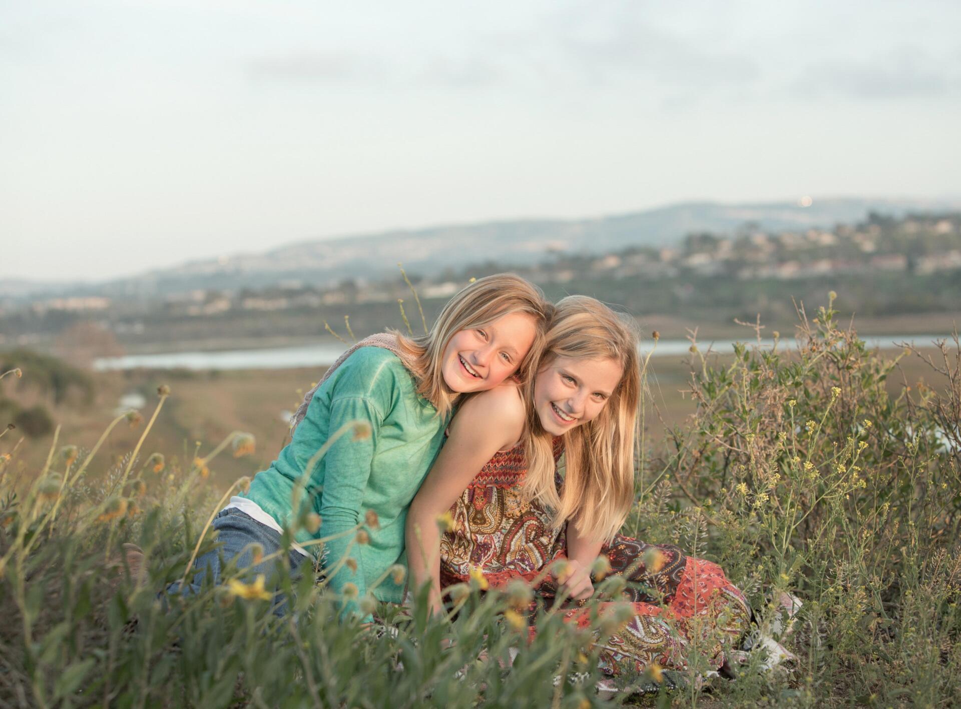 Two young girls are sitting in a field hugging each other.