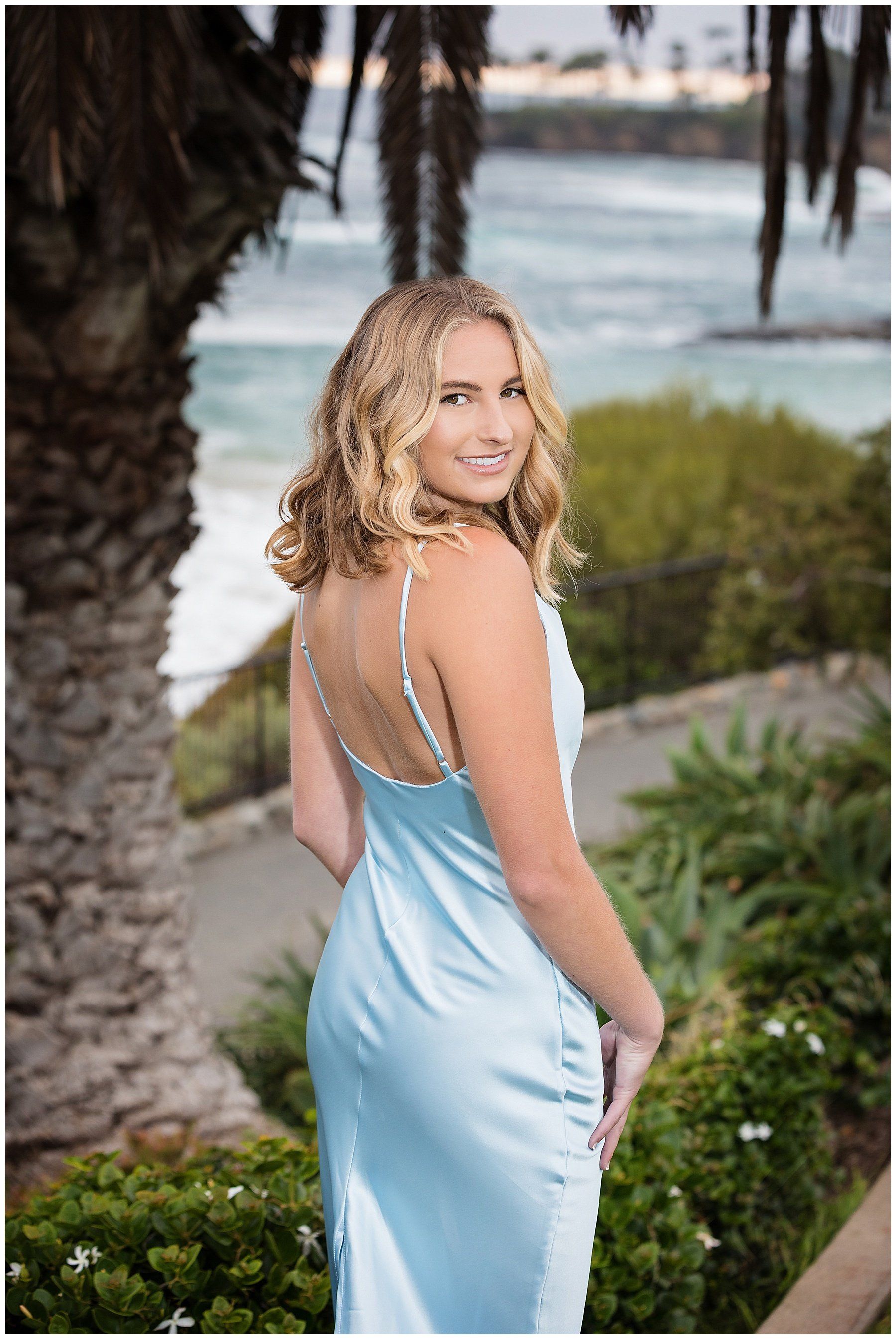 A woman in a light blue dress is standing in front of a palm tree.