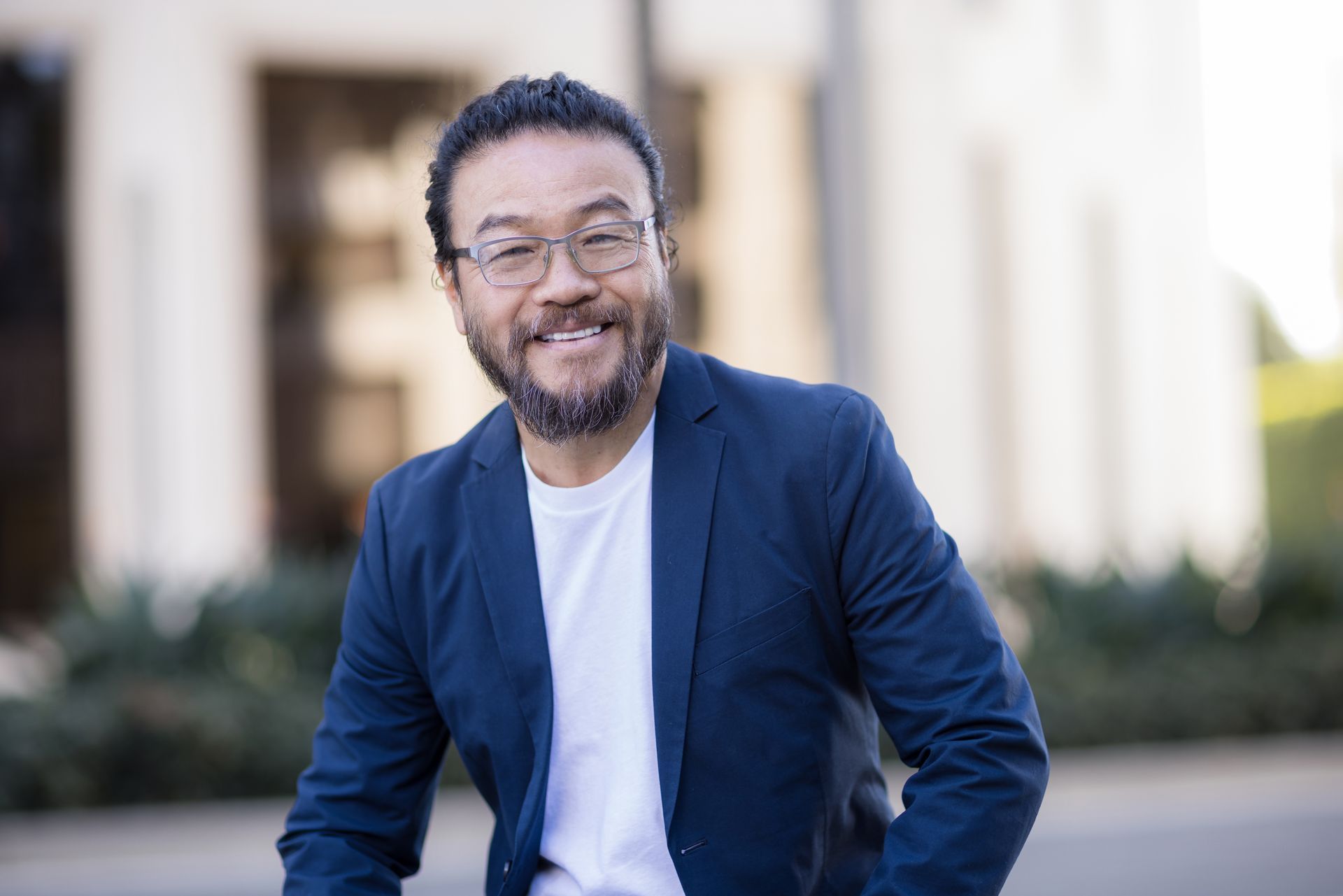 Smiling man in a blue blazer and white shirt seated outdoors with a blurred building background
