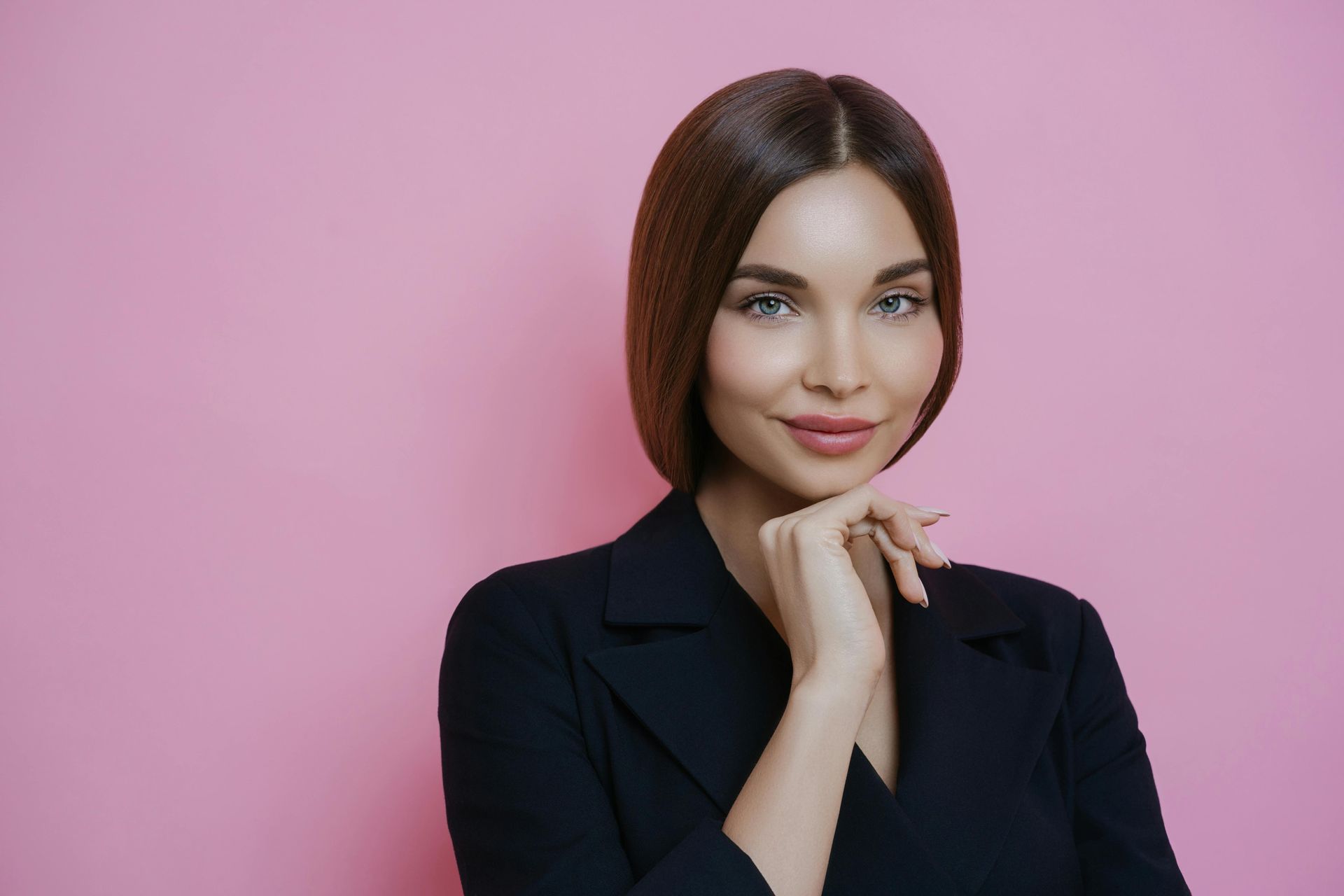 A woman in a black jacket is smiling with her hand on her chin.