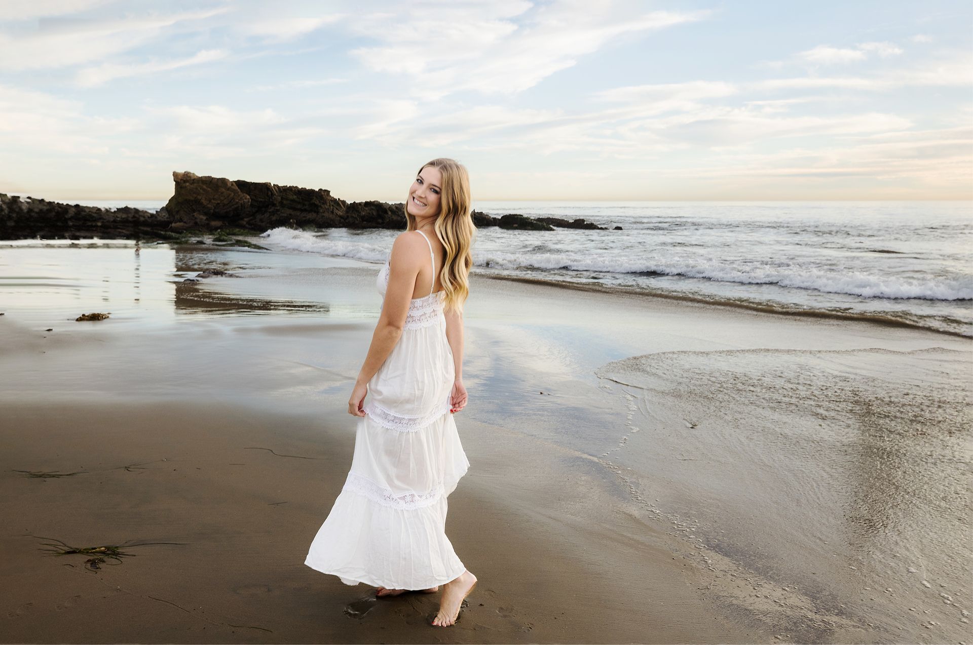 Blond girll in a white dress in front of the ocean at sunset looking over her shoulder.