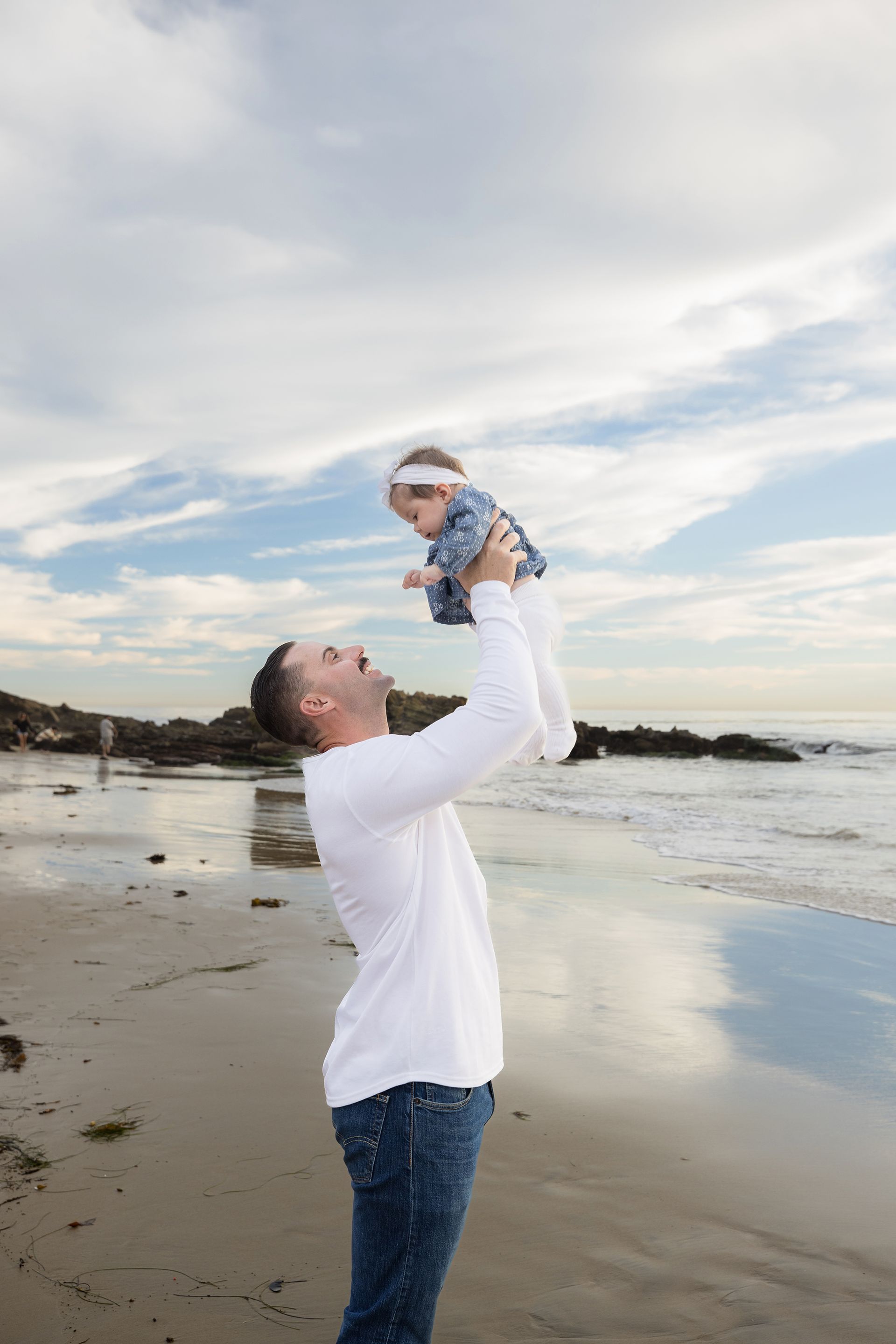 Dad holding his small baby girl up in the air and smiling at her at the beach in Laguna Beach.
