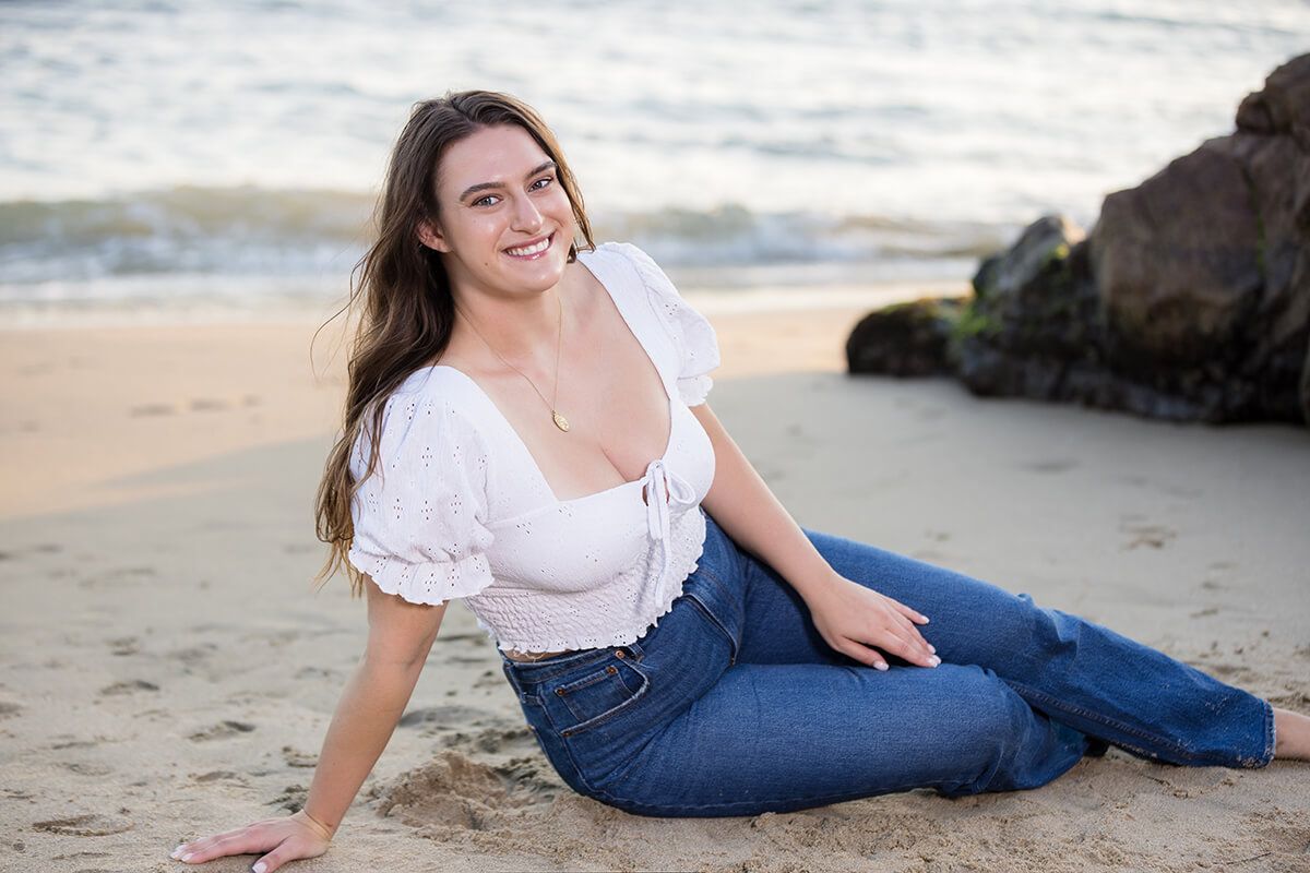 Natural light beach portrait of a young woman sitting on the sand at Picnic Beach in Laguna Beach, wearing a white eyelet blouse and denim jeans