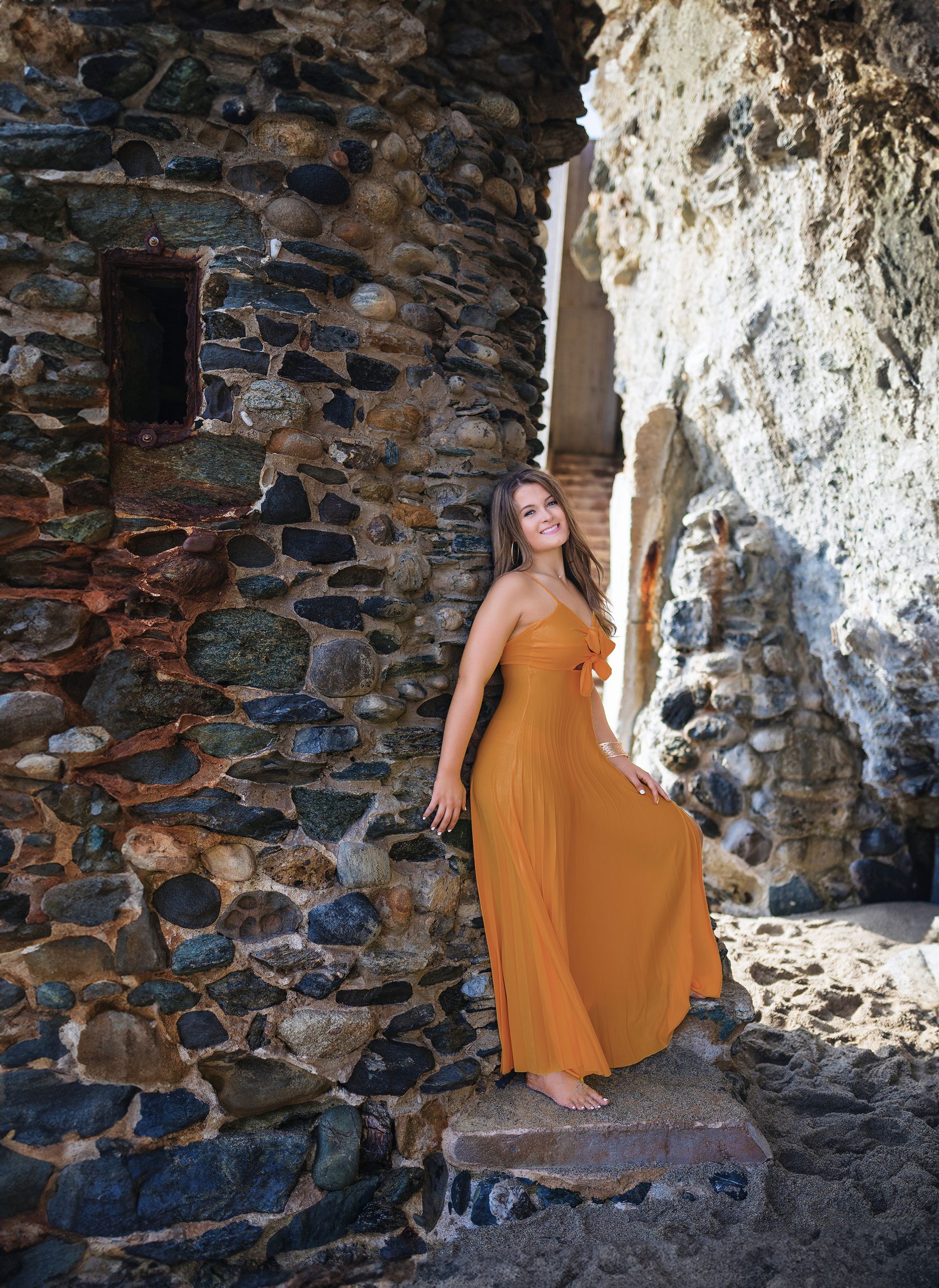 A woman in a long yellow dress is standing in front of a stone wall.
