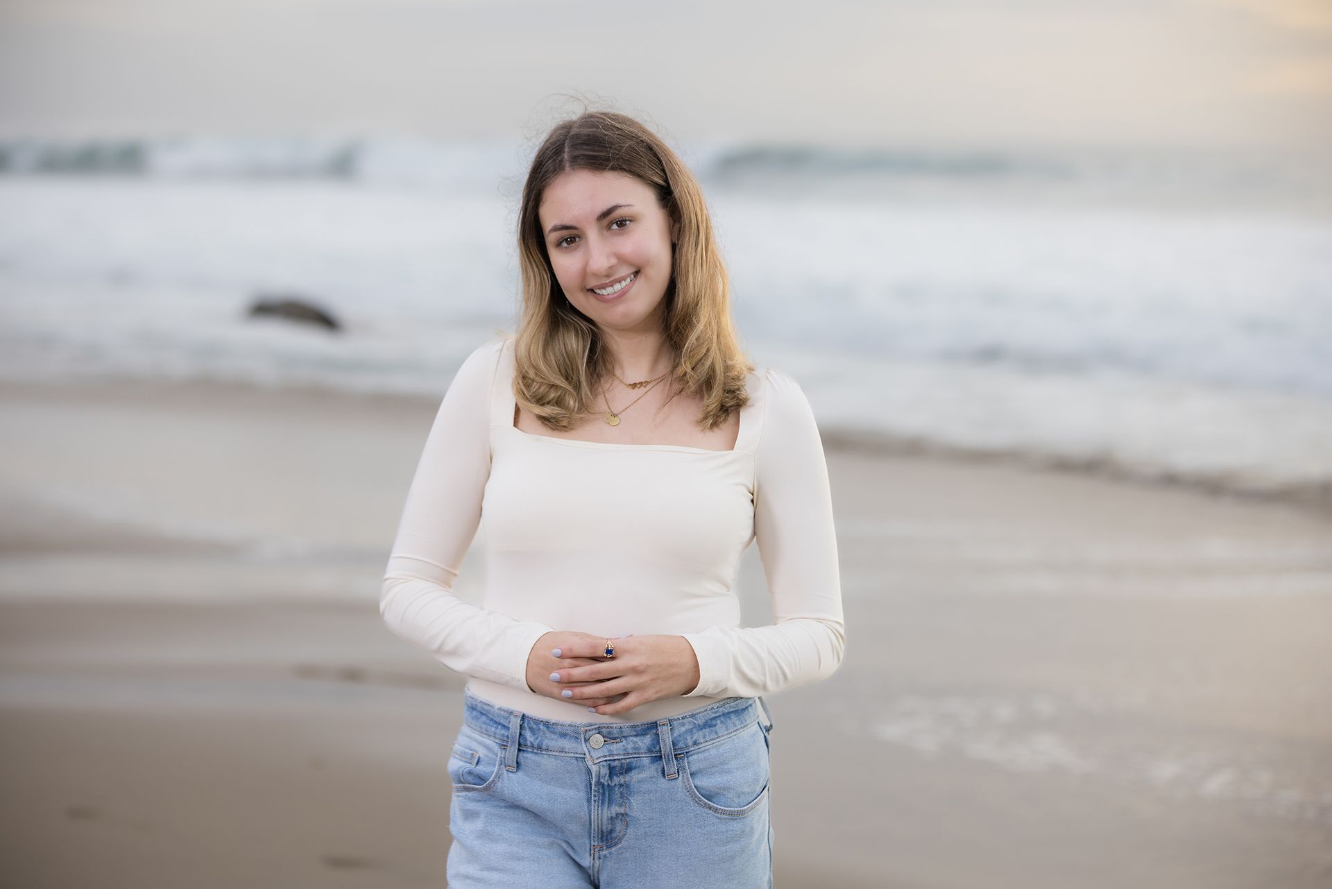 Smiling woman in a white top and jeans standing on a beach with waves behind her