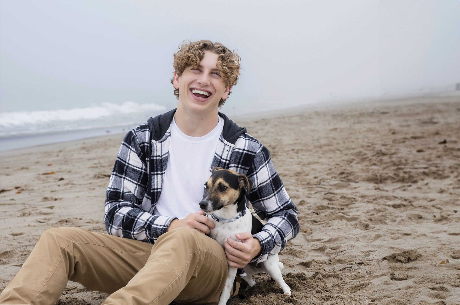 A young man is sitting on the beach with his dog.