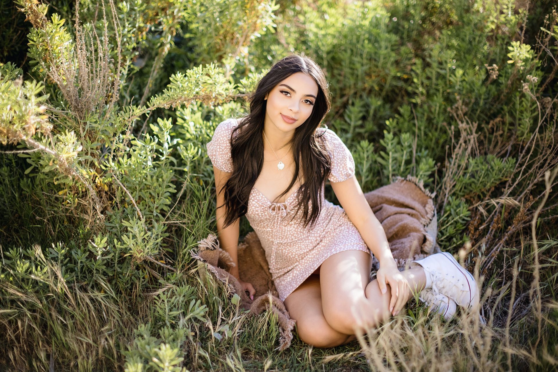 Woman sitting in tall grass and shrubs, wearing a light pink dress and smiling at the camera.