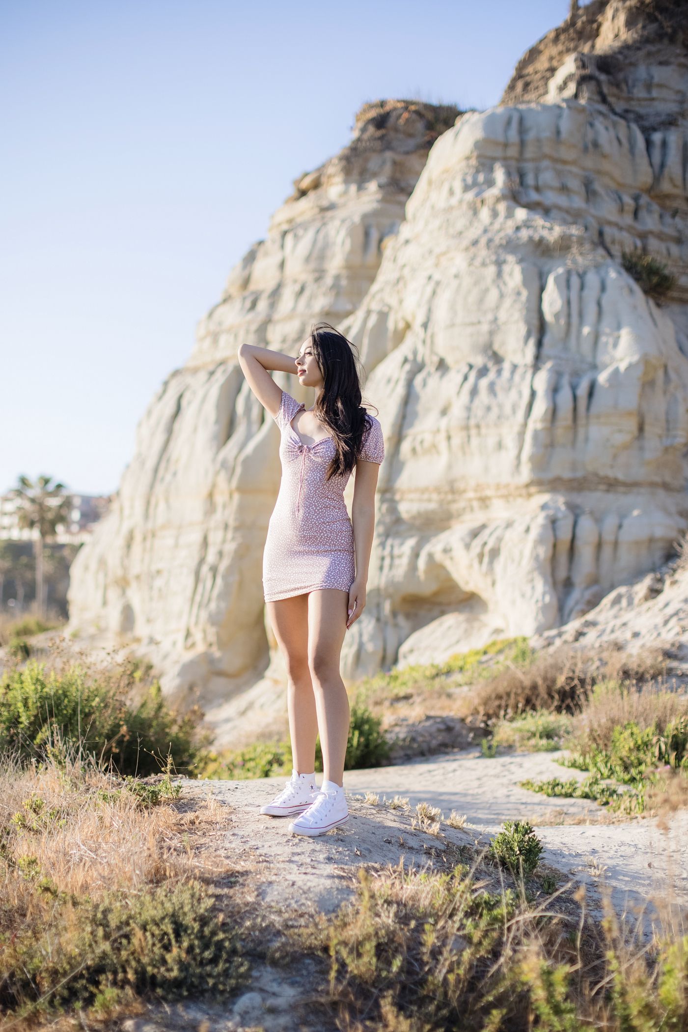 Woman in a pink dress standing on a rocky trail beside a white cliff, reaching toward the sun