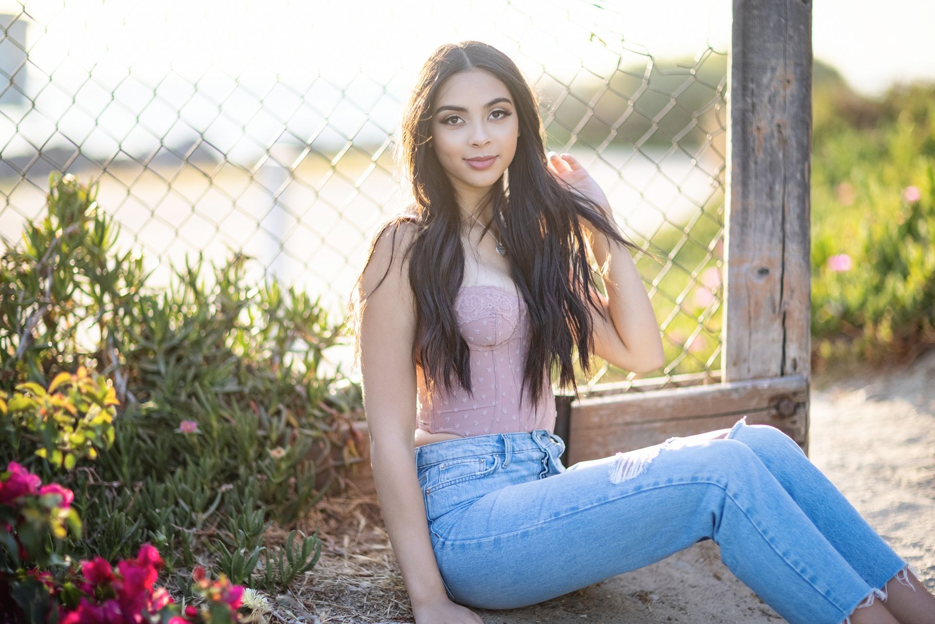 Young woman sitting by a wooden fence in a garden, wearing a pink top and blue jeans, with flowers nearby.