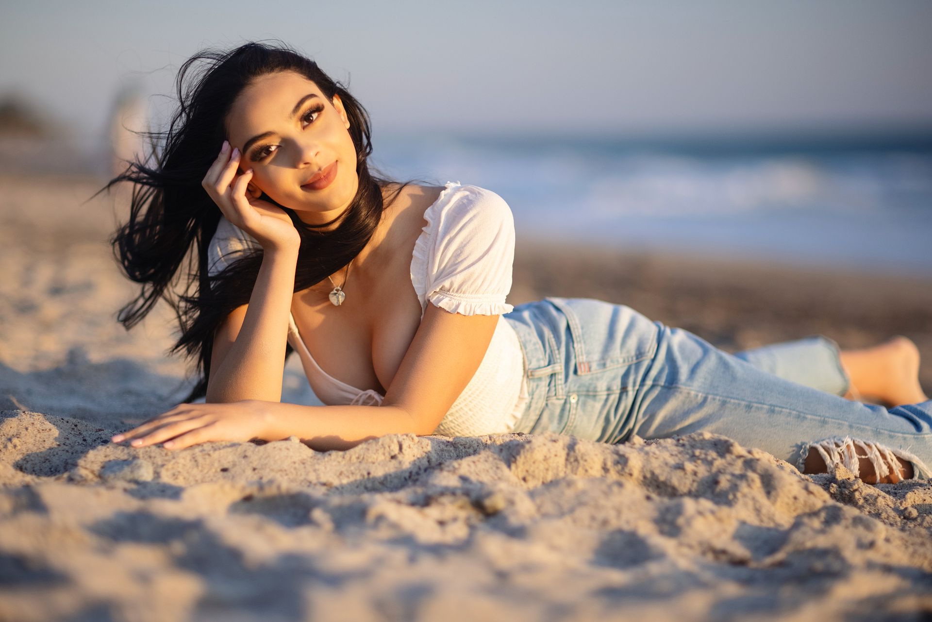 Woman lying on a sandy beach, resting on her elbow and smiling in warm sunlight.